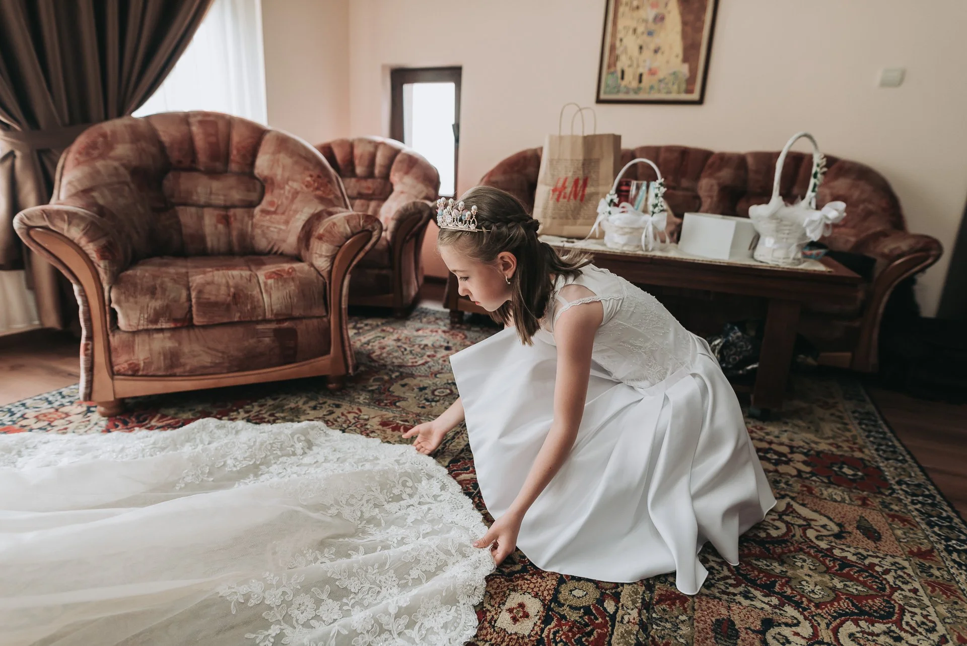 A young girl in a white dress and tiara is kneeling on a patterned rug, adjusting the train of her wedding gown in a living room with brown patterned sofas, a wooden table, and gift bags.
