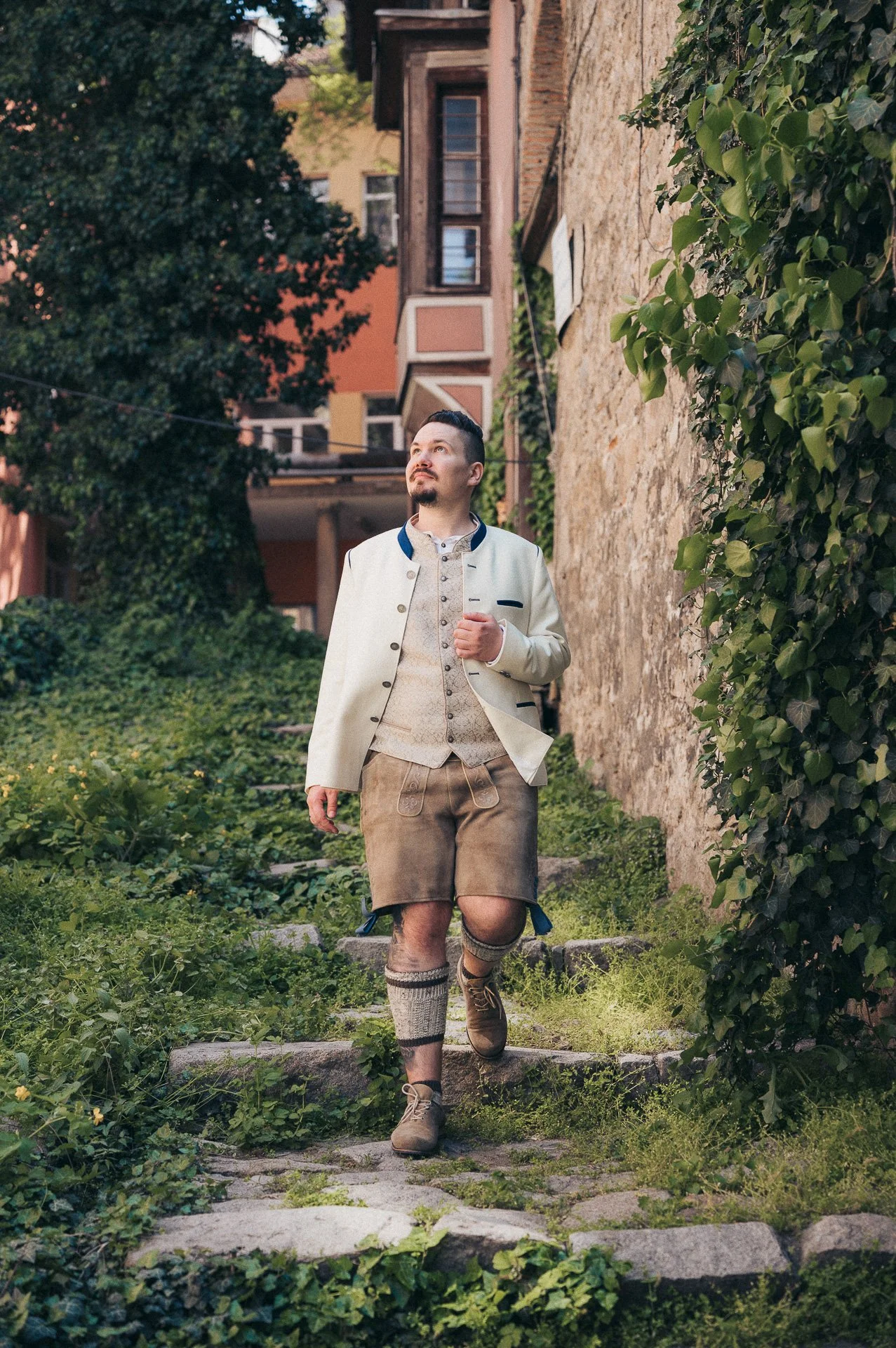 A man dressed in traditional Bavarian clothing, including lederhosen and a knit sock, walking up stone steps outside on a sunny day.