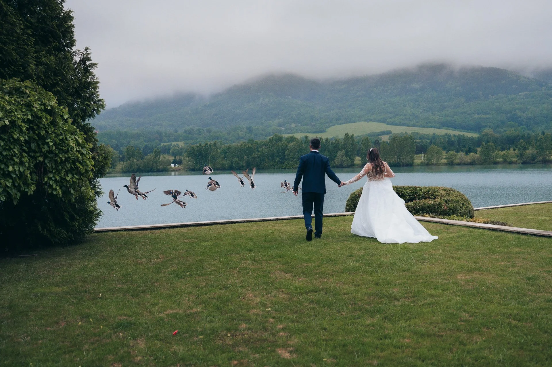 Bride and groom holding hands, walking on grass near a lake, with mountains and mist in the background, and ducks flying nearby.