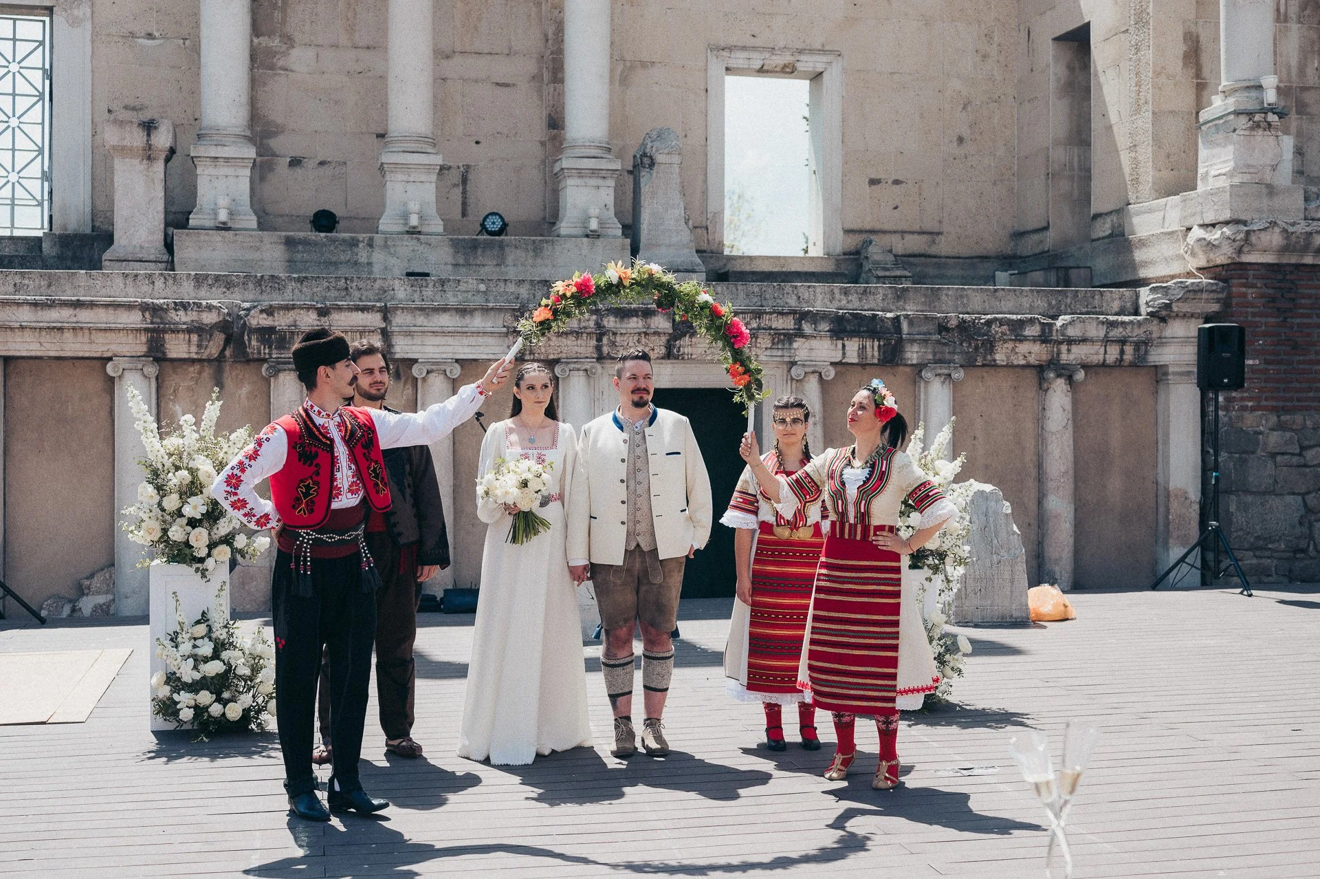 A wedding ceremony with people dressed in traditional Eastern European attire, standing outdoors against a historic stone building backdrop. The couple is surrounded by friends holding a floral arch and bouquets.