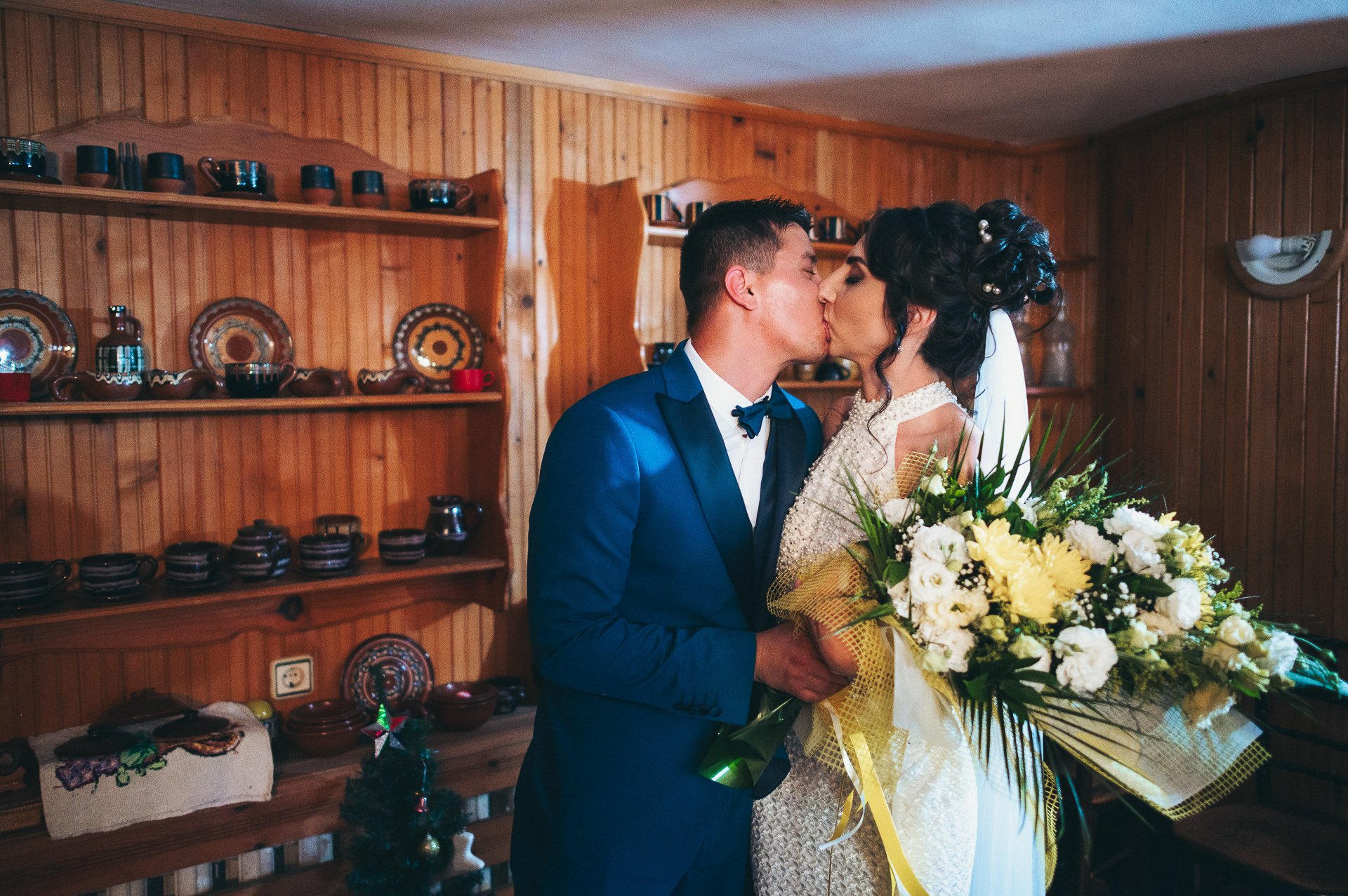 A newlywed couple sharing a kiss, with the groom in a blue suit and bow tie, and the bride in a white wedding dress holding a large bouquet of yellow and white flowers, inside a wooden-paneled room decorated with pottery on shelves.