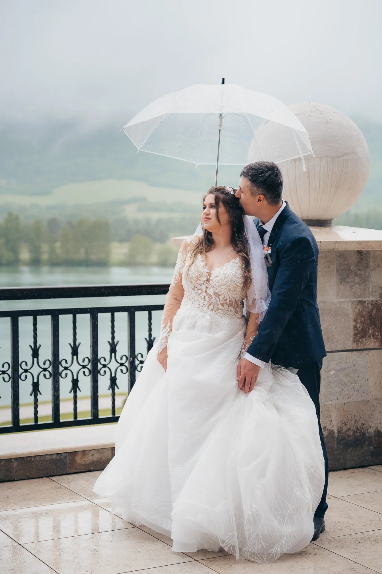 A bride and groom standing under a clear umbrella on a rainy day, with a scenic background of water and green hills.