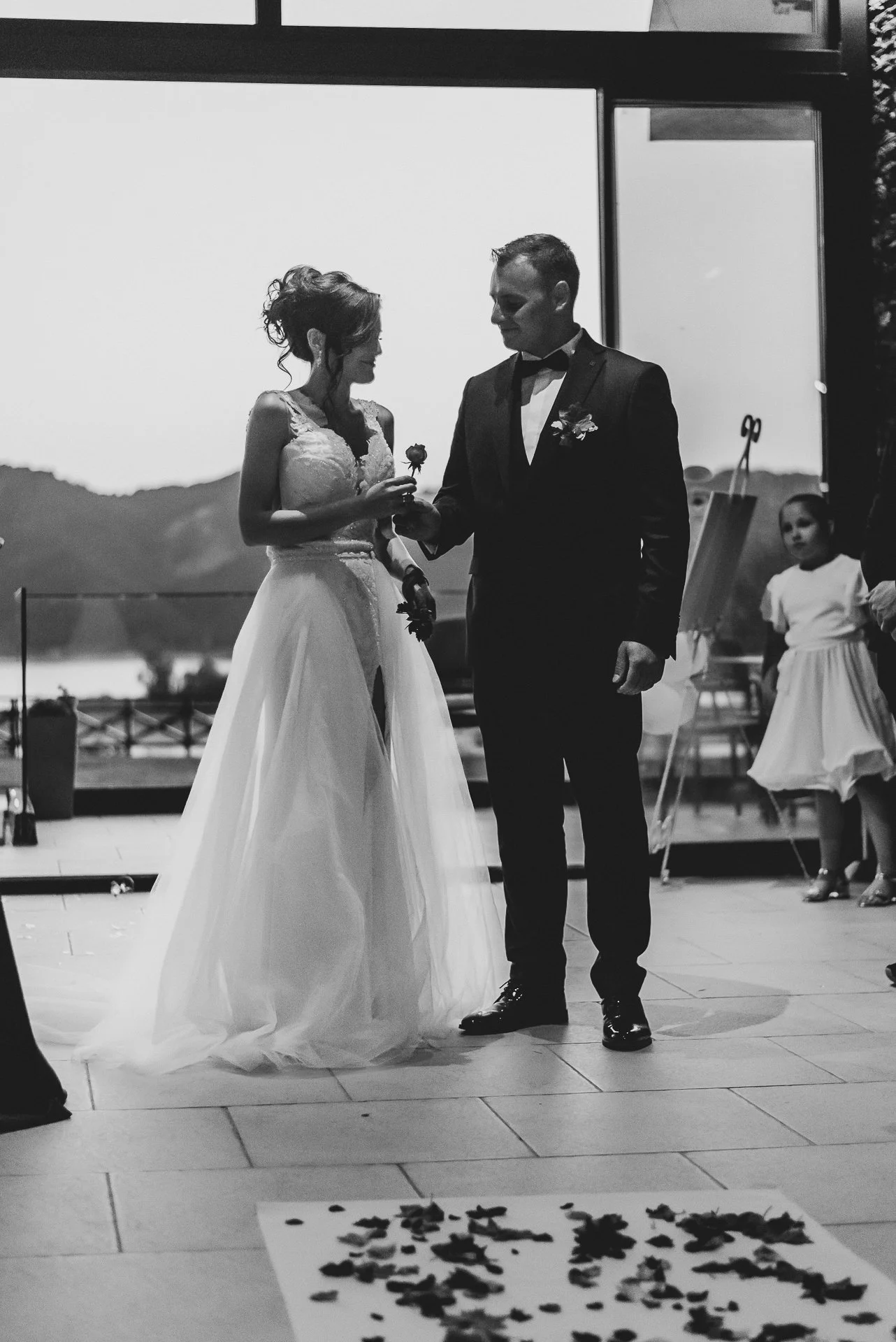 A black and white photo of a bride and groom at their wedding, standing indoors. The bride is holding a flower and the groom is helping her with something. There is a girl standing in the background to the right.