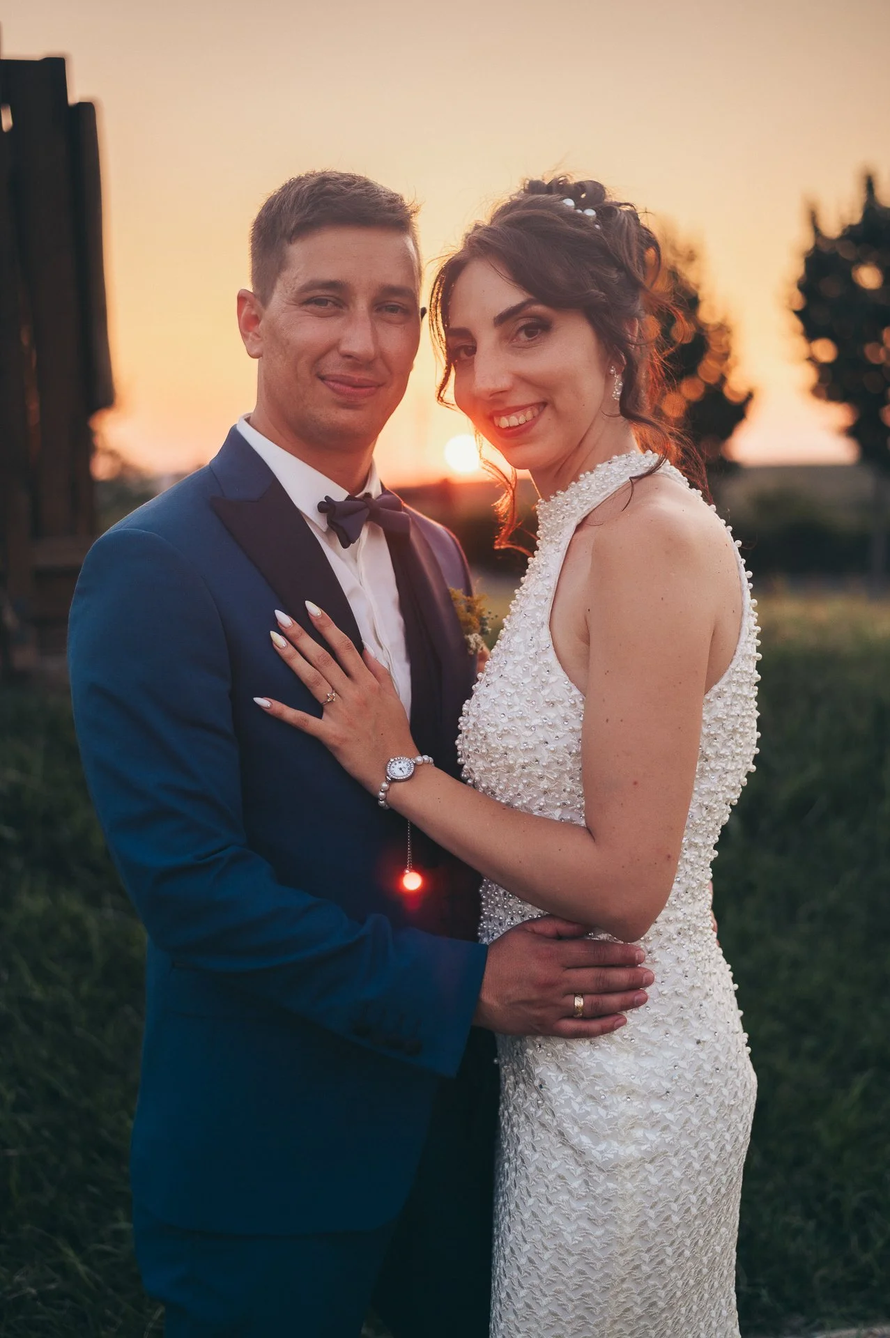 A newlywed couple standing outdoors during sunset, with the man in a blue suit and bow tie and the woman in a white wedding dress, smiling at the camera.