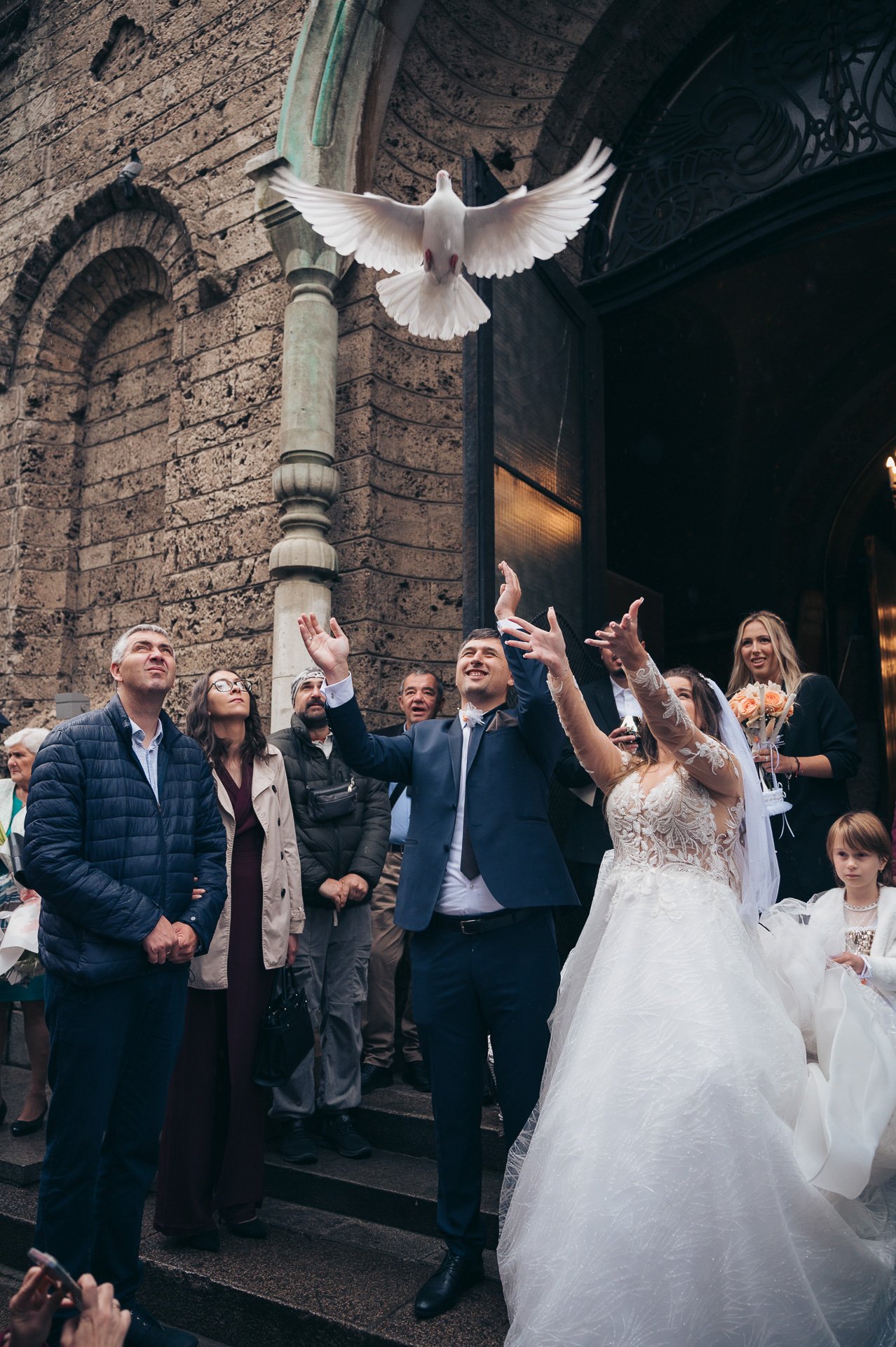 A wedding celebration with a bride and groom releasing a white dove outside a stone building.