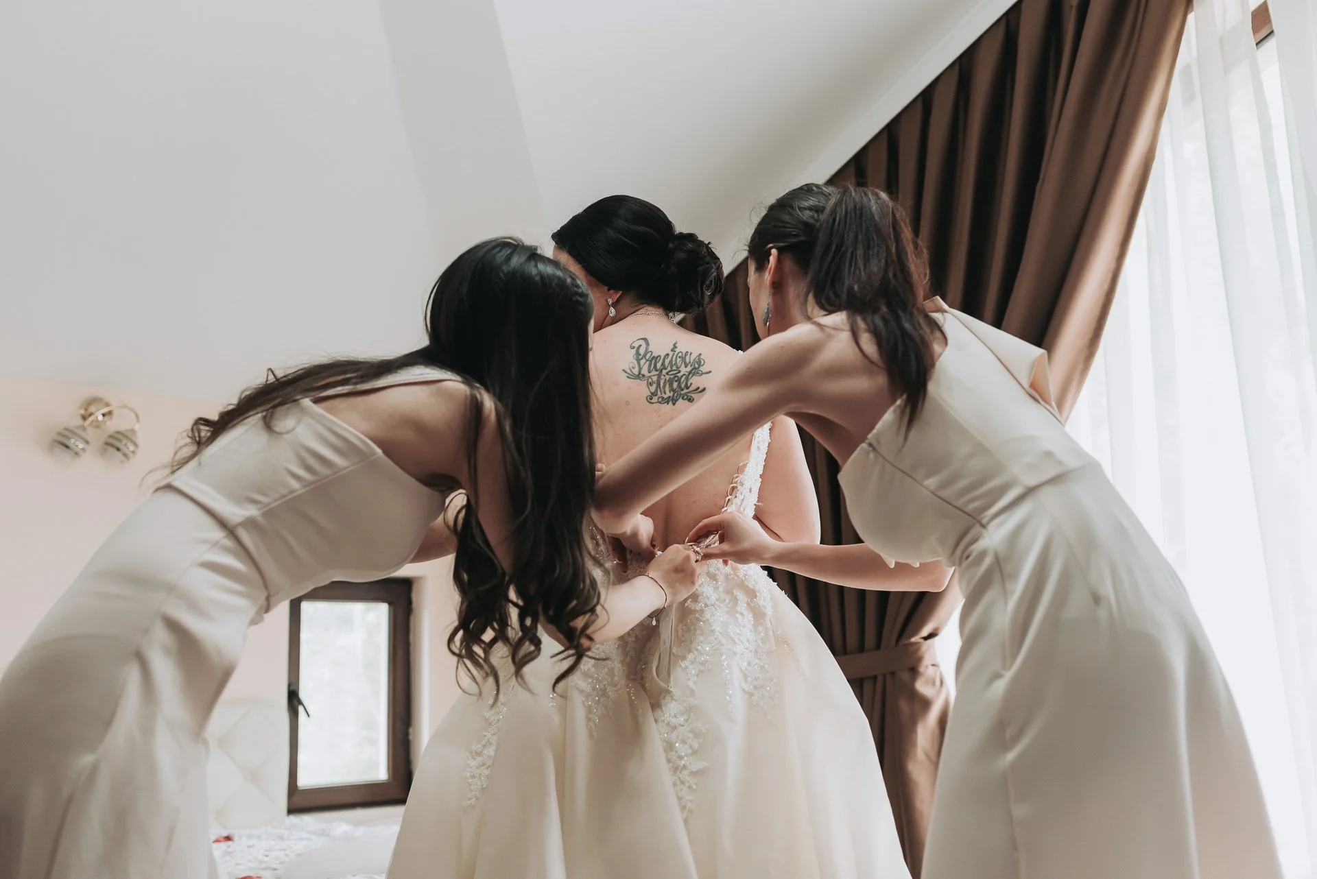 Three women helping a bride with her wedding dress, near a window with brown curtains.