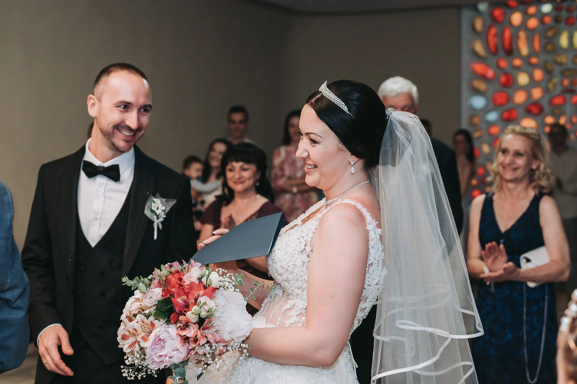 Bride and groom exchanging vows during wedding ceremony, surrounded by guests. The bride holds a bouquet and wears a lace wedding gown and veil, while the groom wears a black tuxedo with a bow tie.