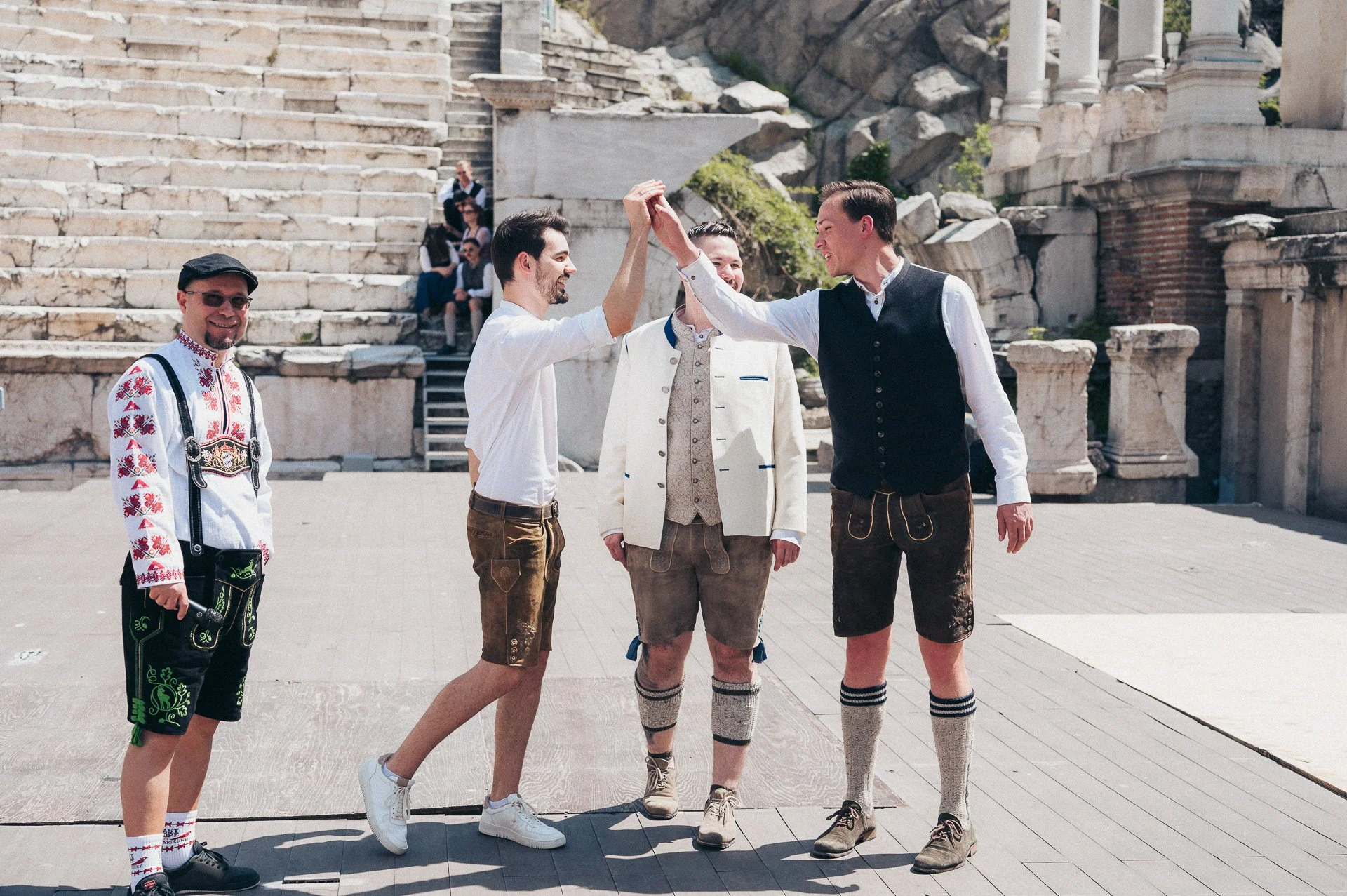 Four men dressed in traditional Bavarian attire, standing on a stone platform in front of ancient ruins, sharing a high-five, with other people seated on stairs in the background.
