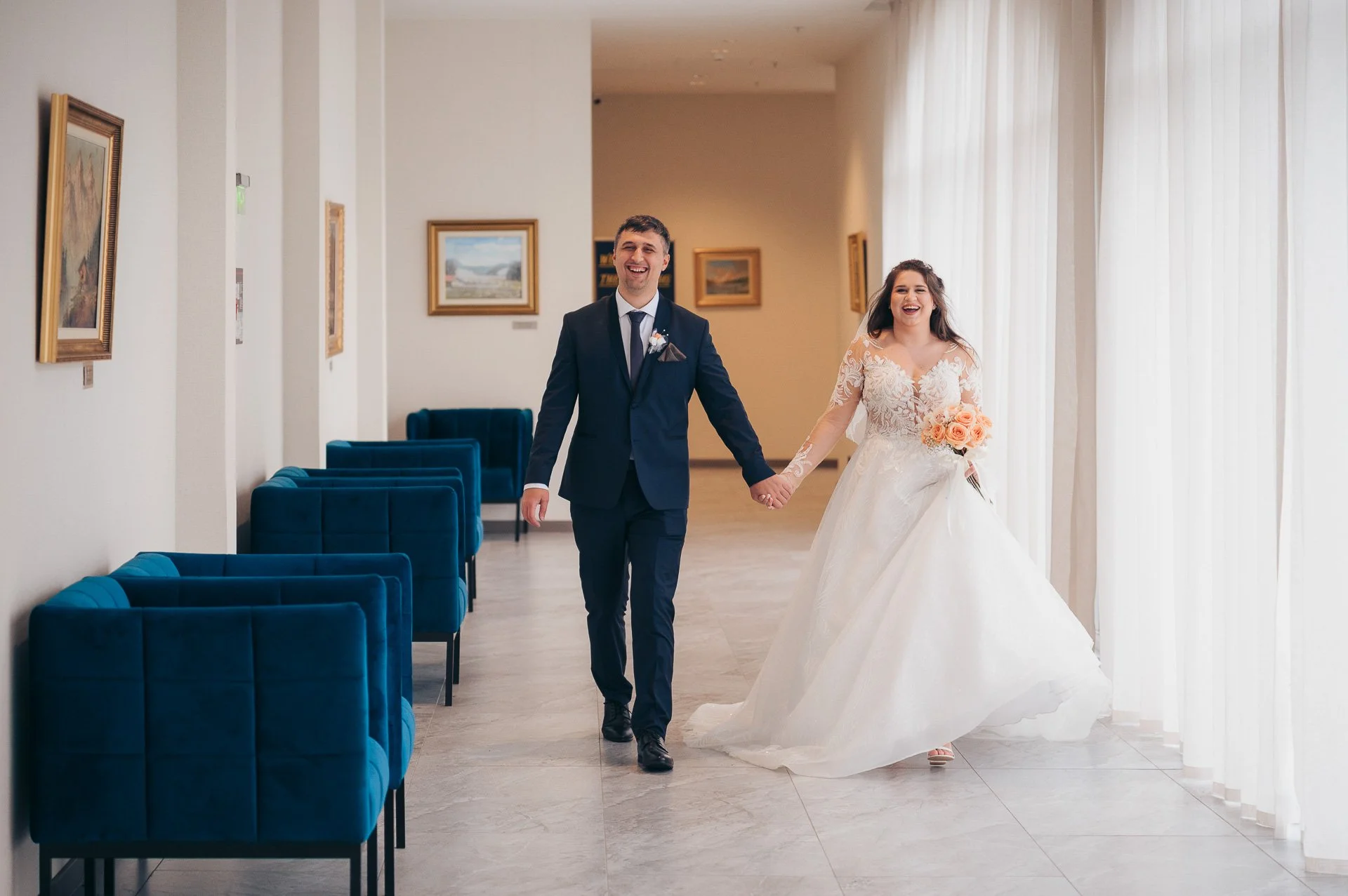 A newlywed couple in wedding attire walking hand-in-hand through an art gallery, smiling and looking joyful.