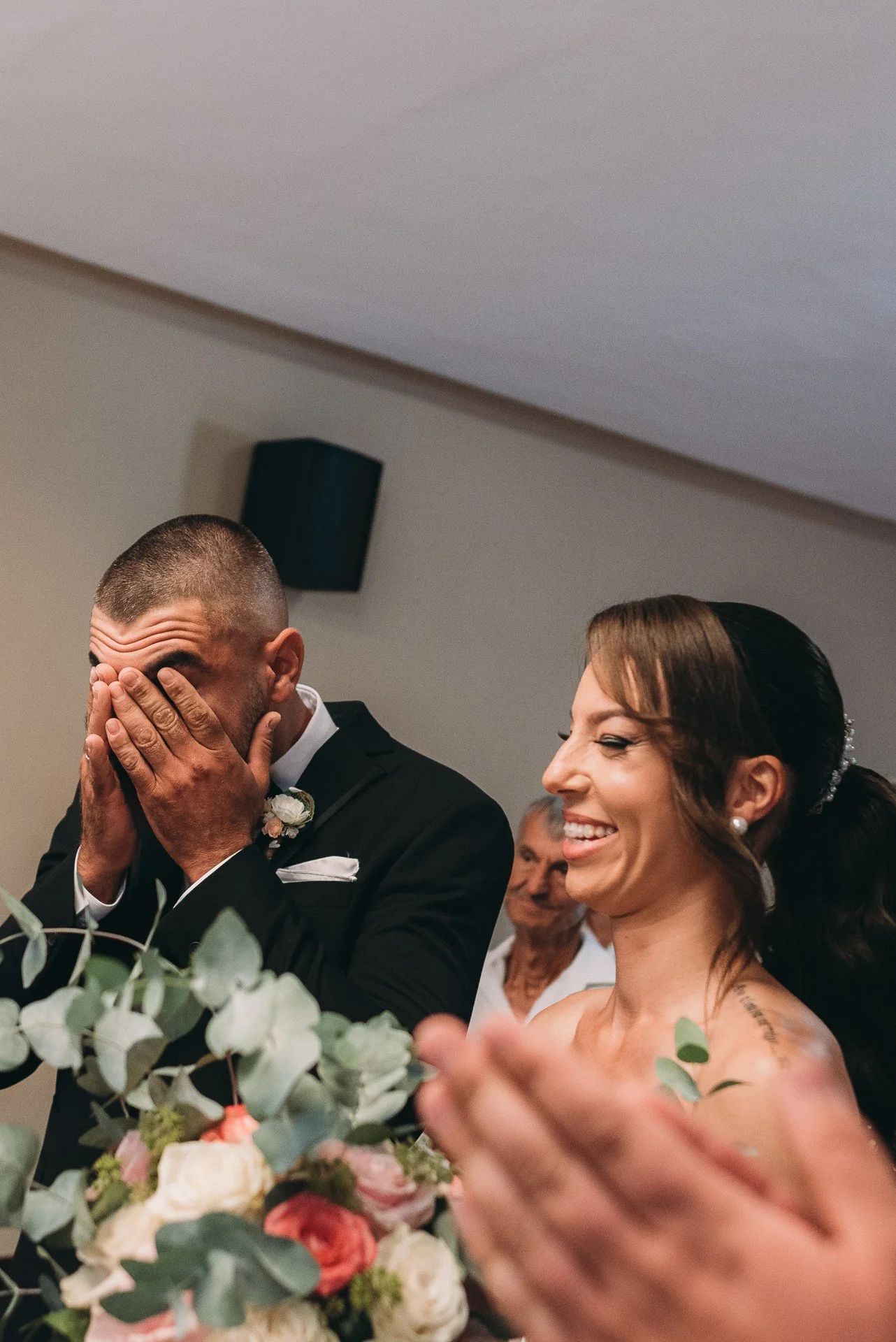 A groom covering his face with his hands and a smiling bride with dark hair tied back, wearing pearl earrings, at a wedding reception with flowers in the foreground and a guest in the background