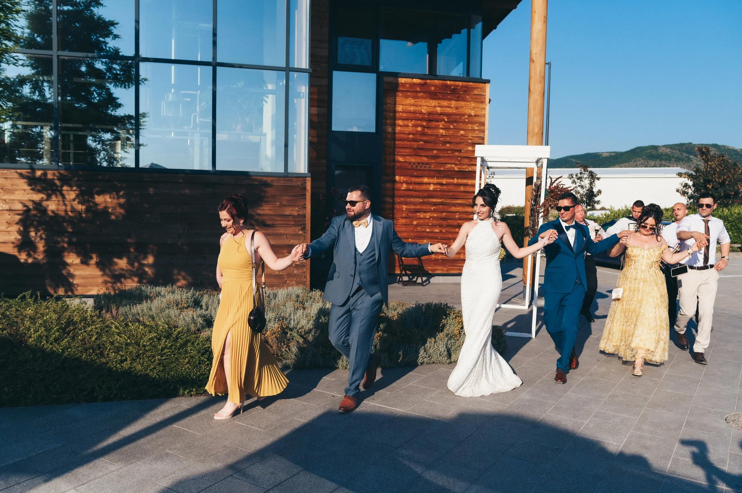 A group of people dressed in formal attire holding hands and dancing outdoors near a modern building with wooden and glass elements.