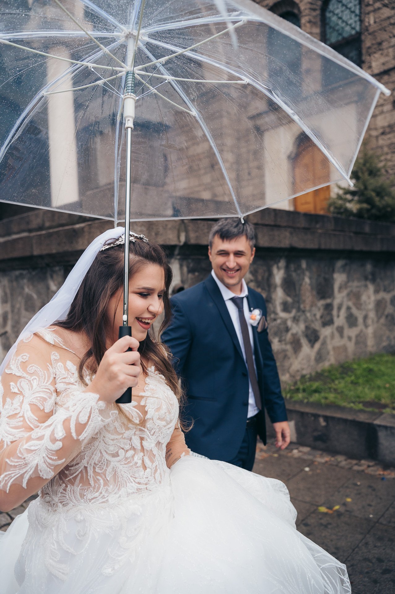 A newlywed couple smiles under a clear umbrella outside on a rainy day. The bride wears a lace wedding dress and veil, and the groom is in a dark suit with a white shirt and tie.