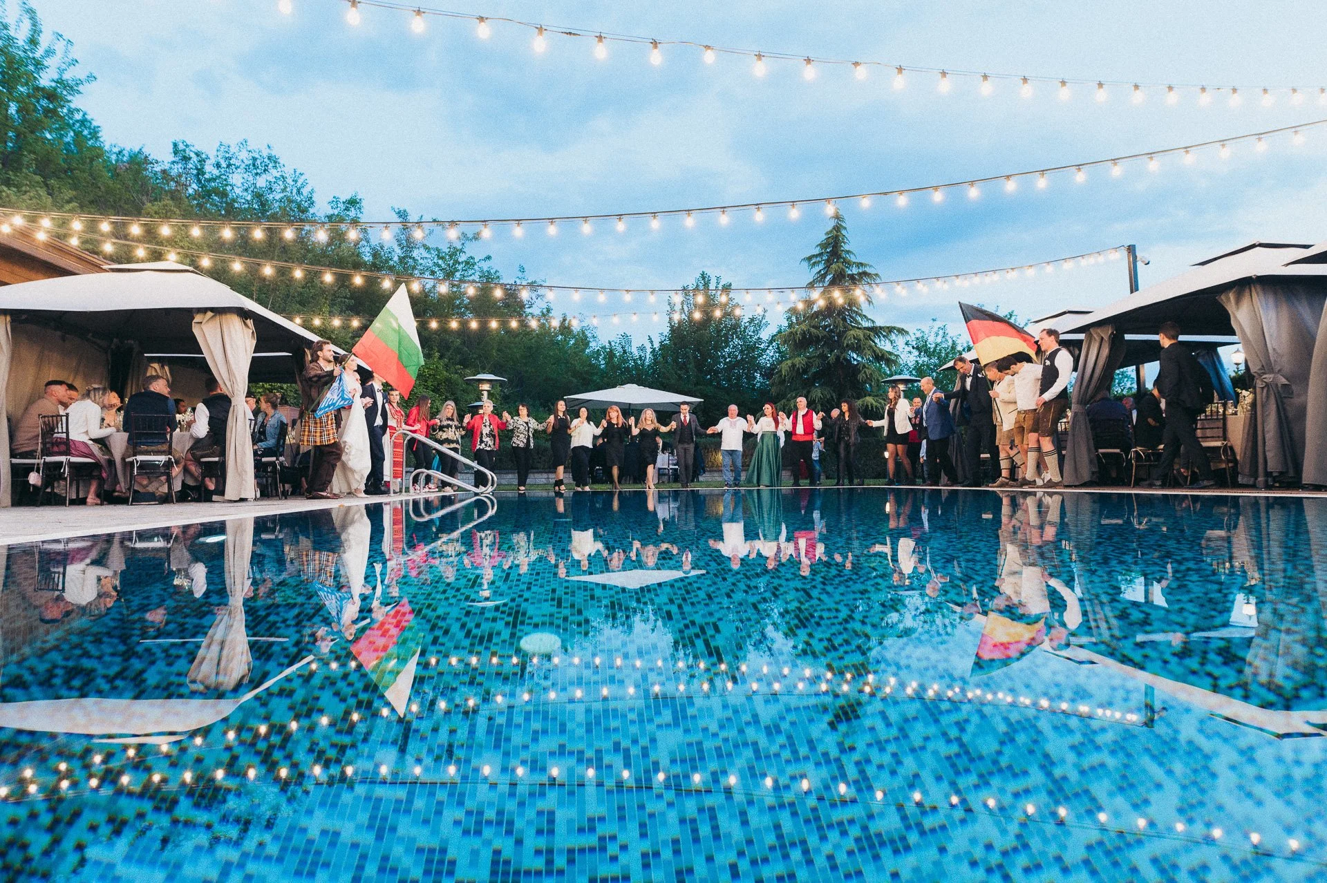 People dancing in a circle by a swimming pool at an outdoor evening party with string lights overhead, tents on the sides, and some guests seated.