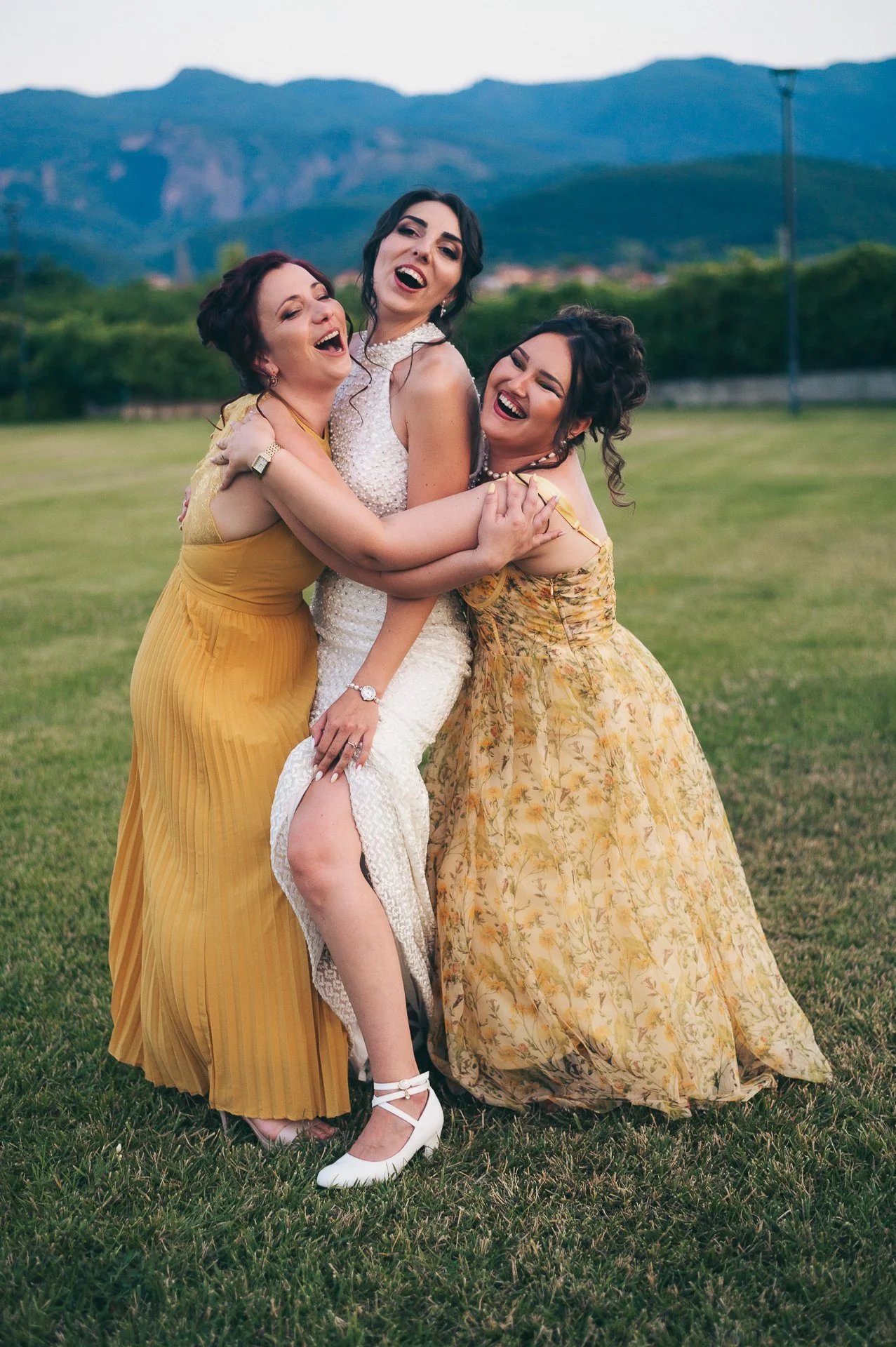 Three women in dresses hugging and smiling outdoors with hills in the background.