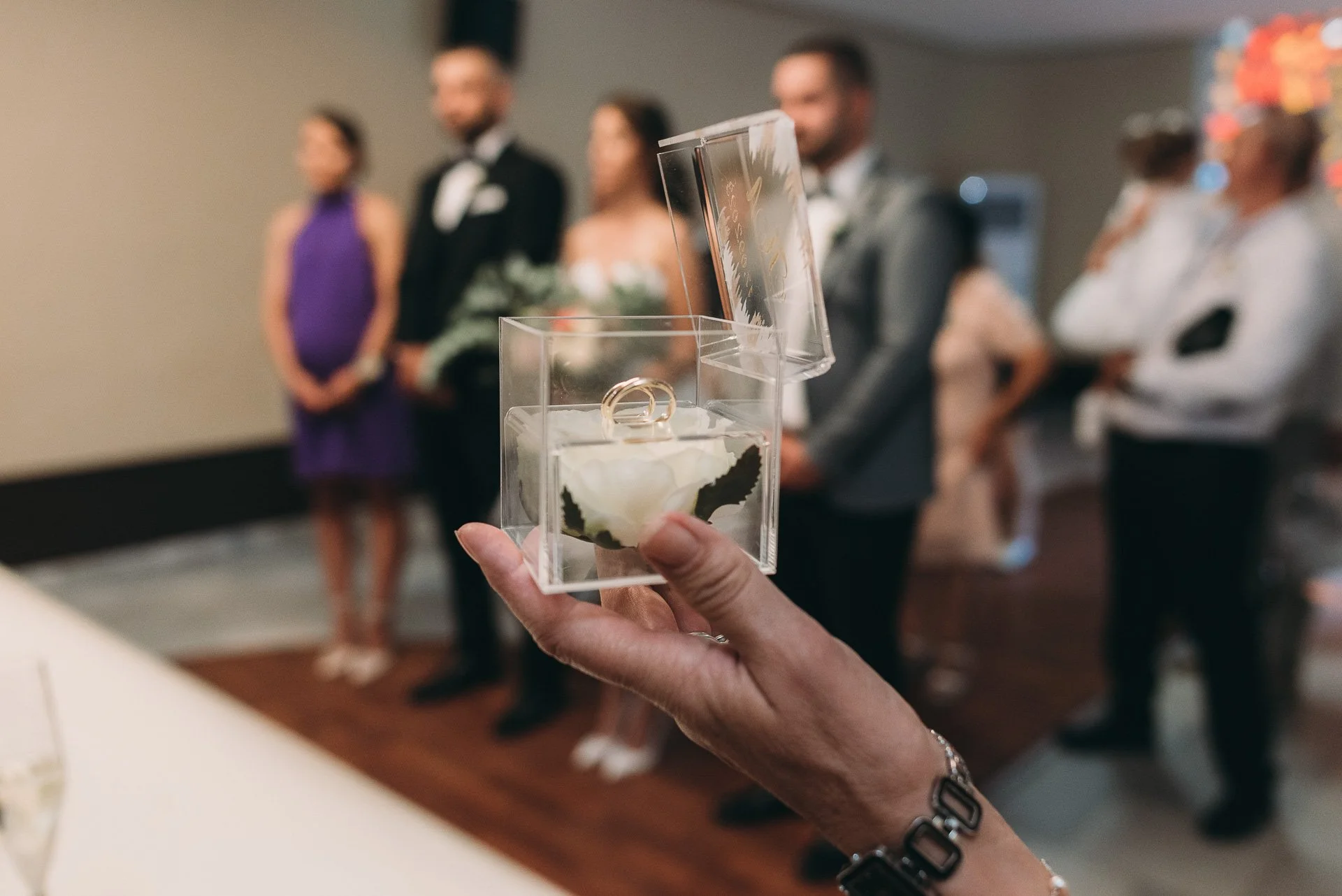 A hand holding a small transparent box with wedding rings and a flower inside, with a blurred wedding ceremony in the background.