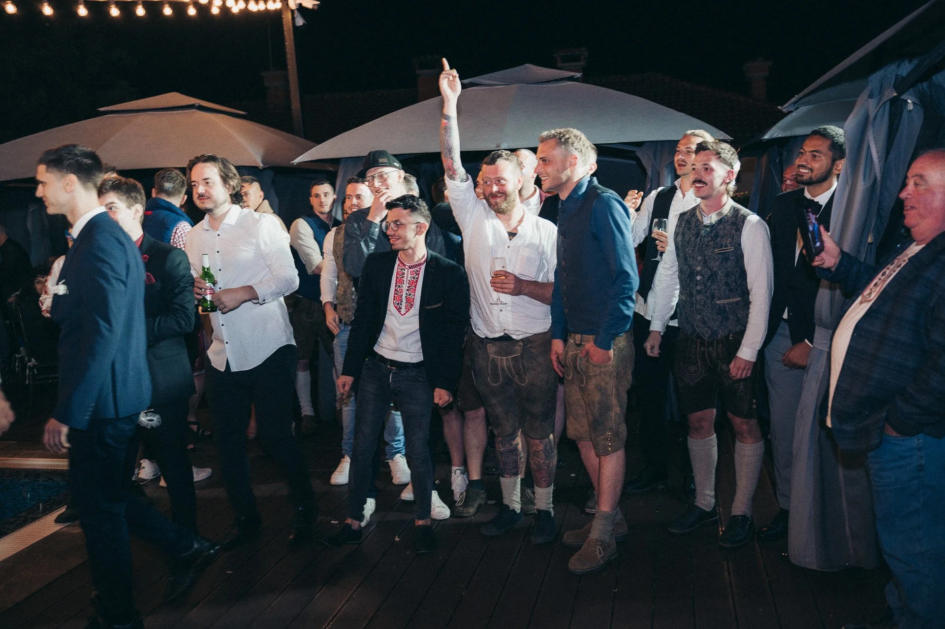 Group of people at an outdoor evening event, with some wearing traditional attire, under umbrellas, enjoying drinks, dancing, and socializing.
