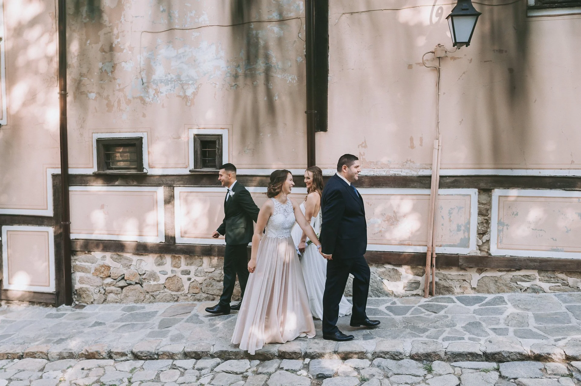 A wedding party with two couples walking on a stone sidewalk in front of an old, weathered building with pink and beige walls and small upper windows, during daytime.