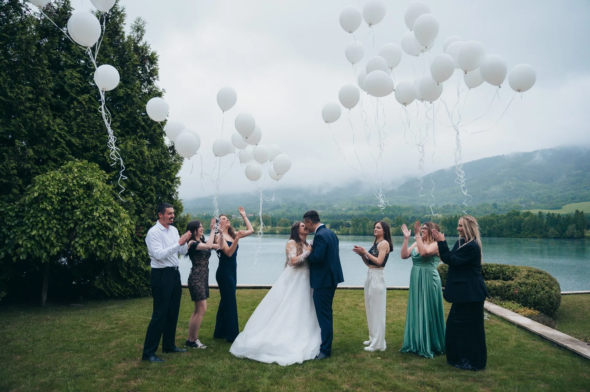 Wedding celebration outdoors with a bride and groom kissing amid friends. White balloons in the sky by a lake with mountains in the background.