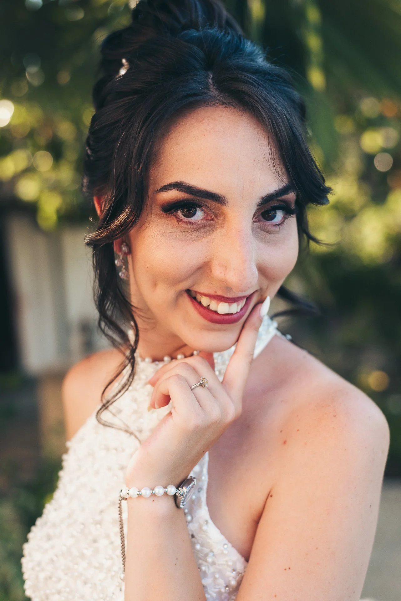 Close-up of a smiling woman with dark hair, wearing a pearl necklace and earrings, with her finger near her lips, outdoors with blurred green foliage in the background.