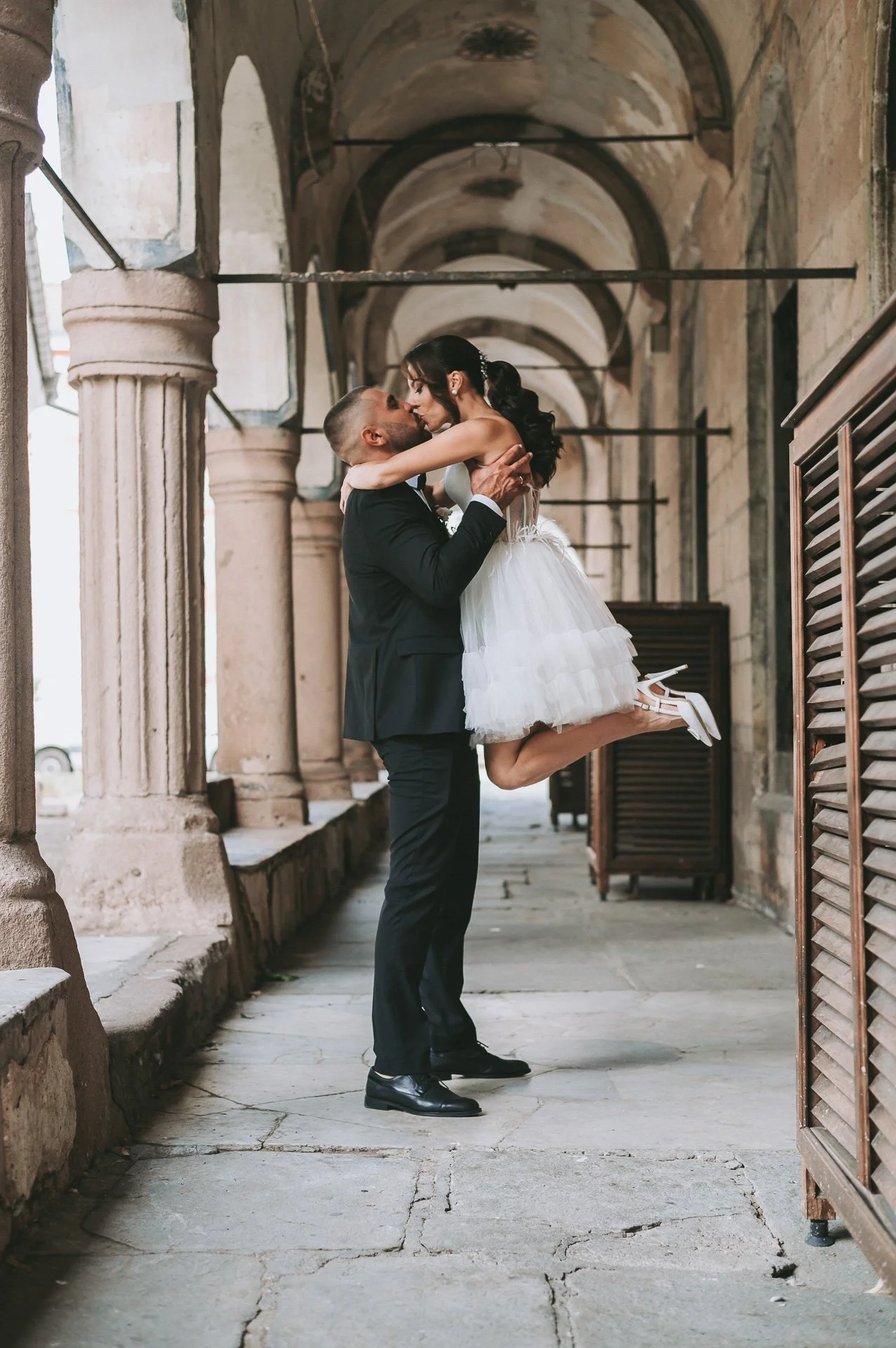 A couple in wedding attire, with the groom lifting the bride in his arms, sharing a kiss in a historic, arched corridor with stone columns and vaulted ceilings.