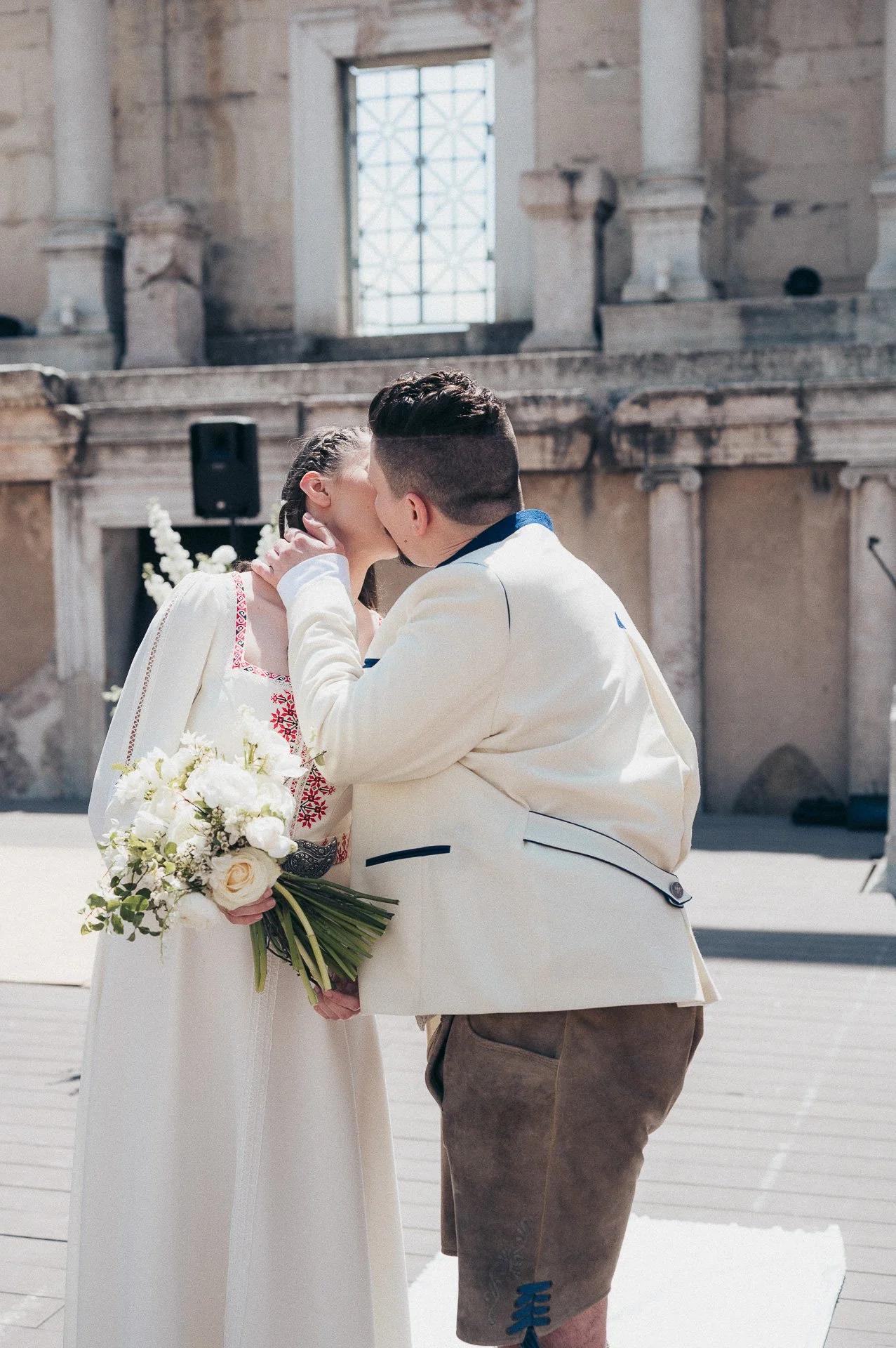 A couple getting married, sharing a kiss during their wedding ceremony, with the bride holding a bouquet of white flowers, in a historic stone building.