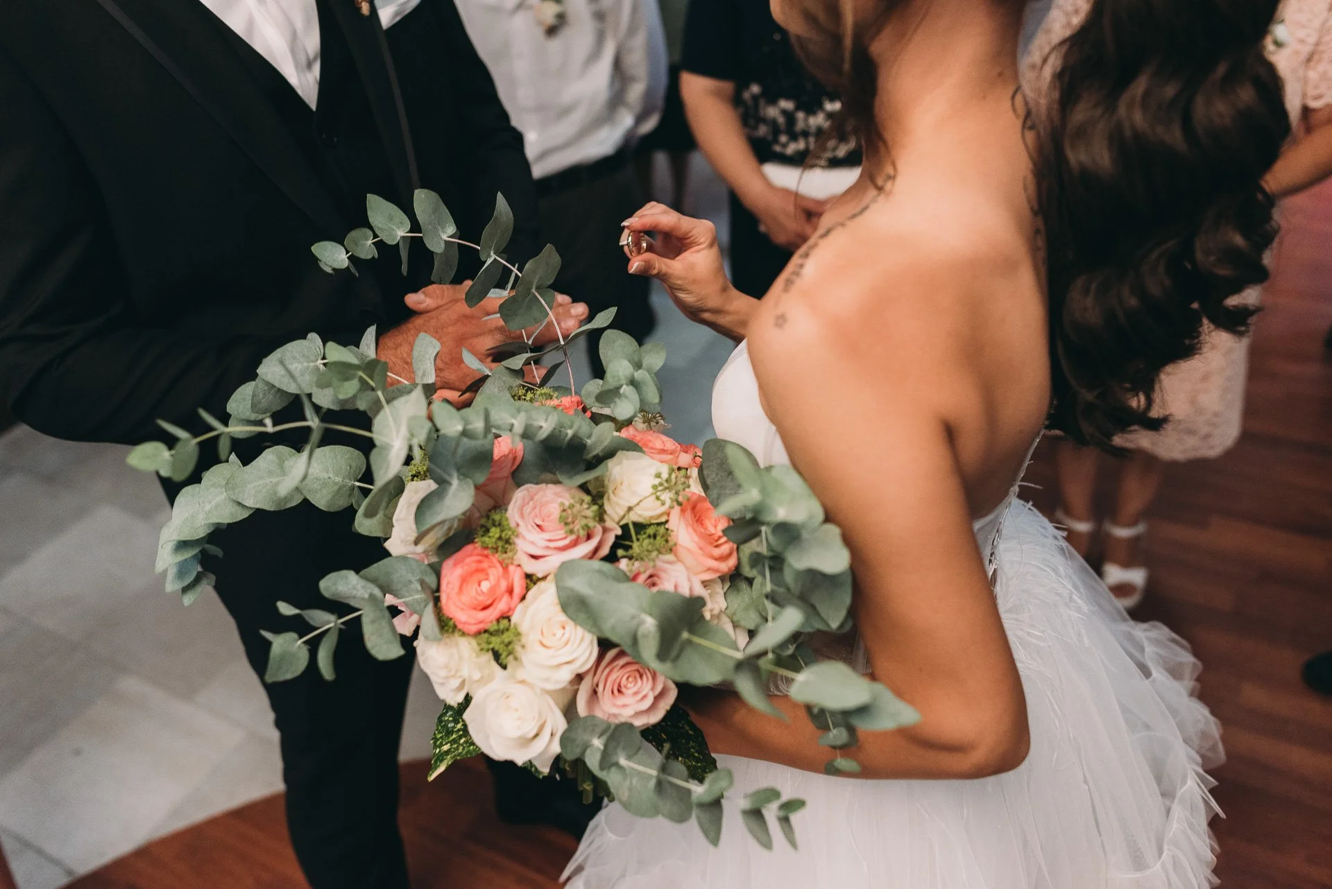 A bride with long dark curly hair in a white wedding dress is holding a bouquet of pink and white roses with green leaves, while appearing to exchange rings with a person dressed in black. There are other people in the background.