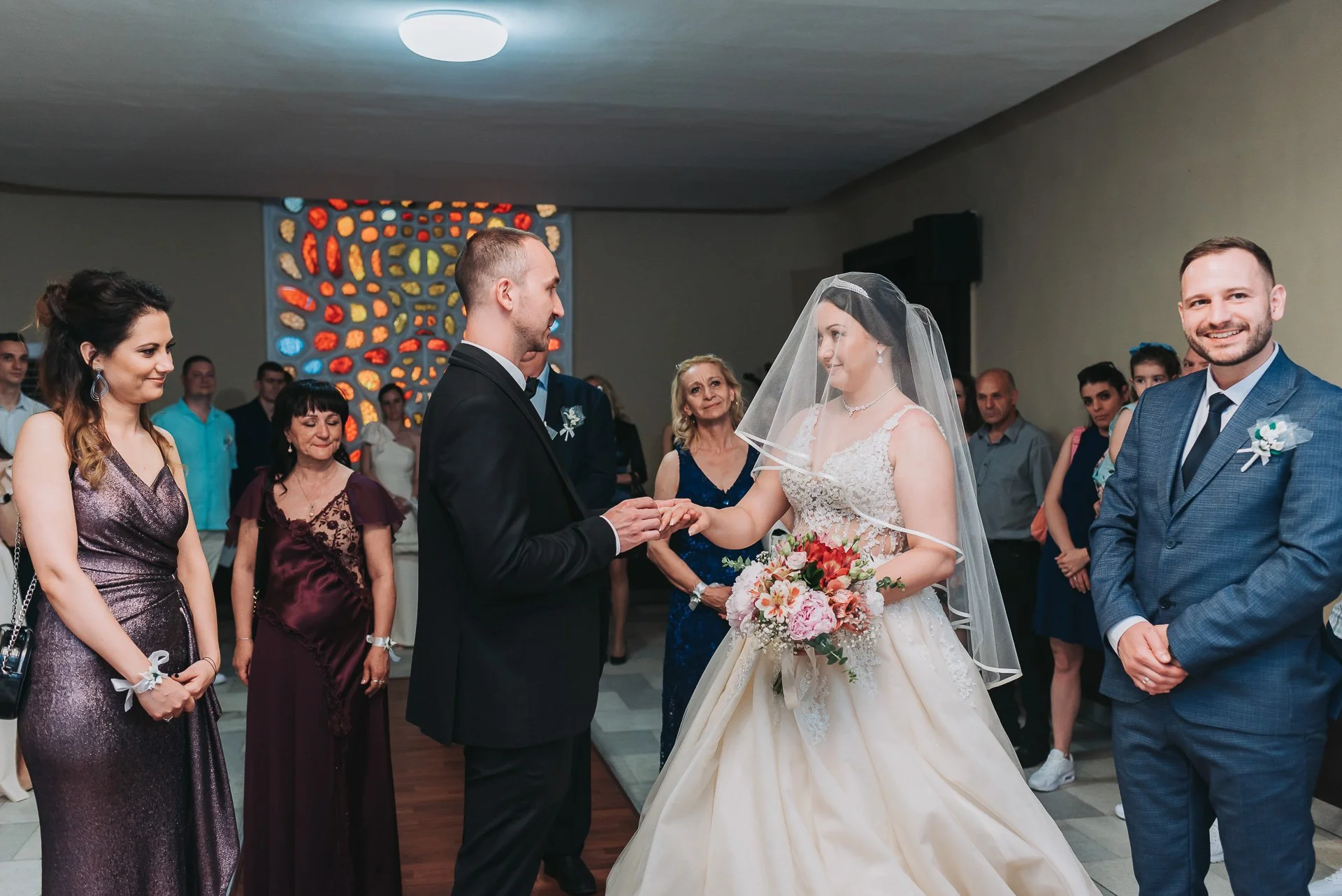Wedding ceremony with bride and groom exchanging rings surrounded by family and friends in an indoor venue.