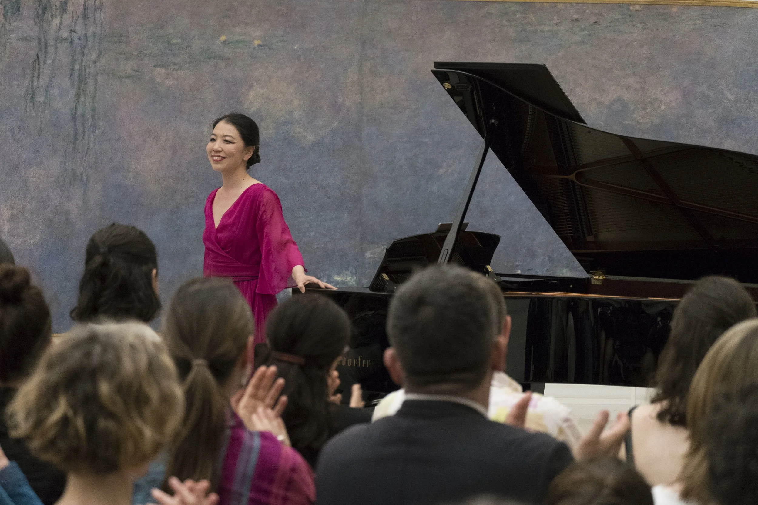 Une femme en robe rose se tient à côté d'un piano à queue noir dans une salle de concert, saluant ou remerciant le public. Le fond est une toile de couleur sombre.