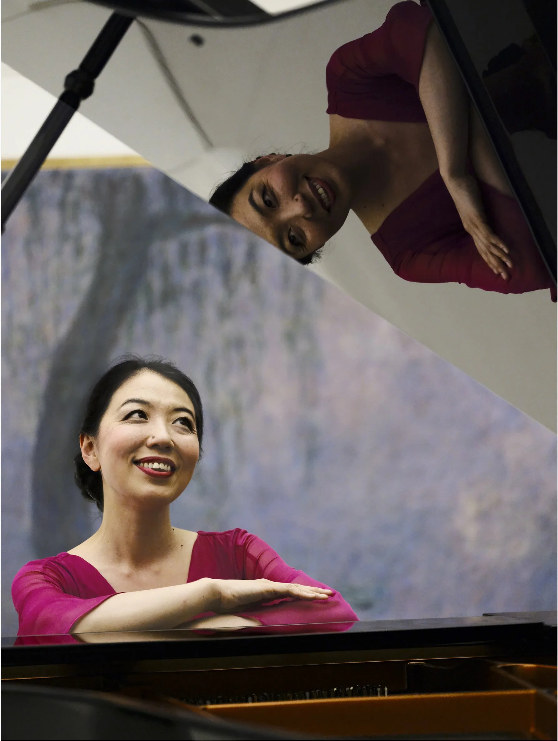 Une femme souriante joue du piano avec ses bras croisés, regardant vers la droite, avec un décor de paysage en arrière-plan.