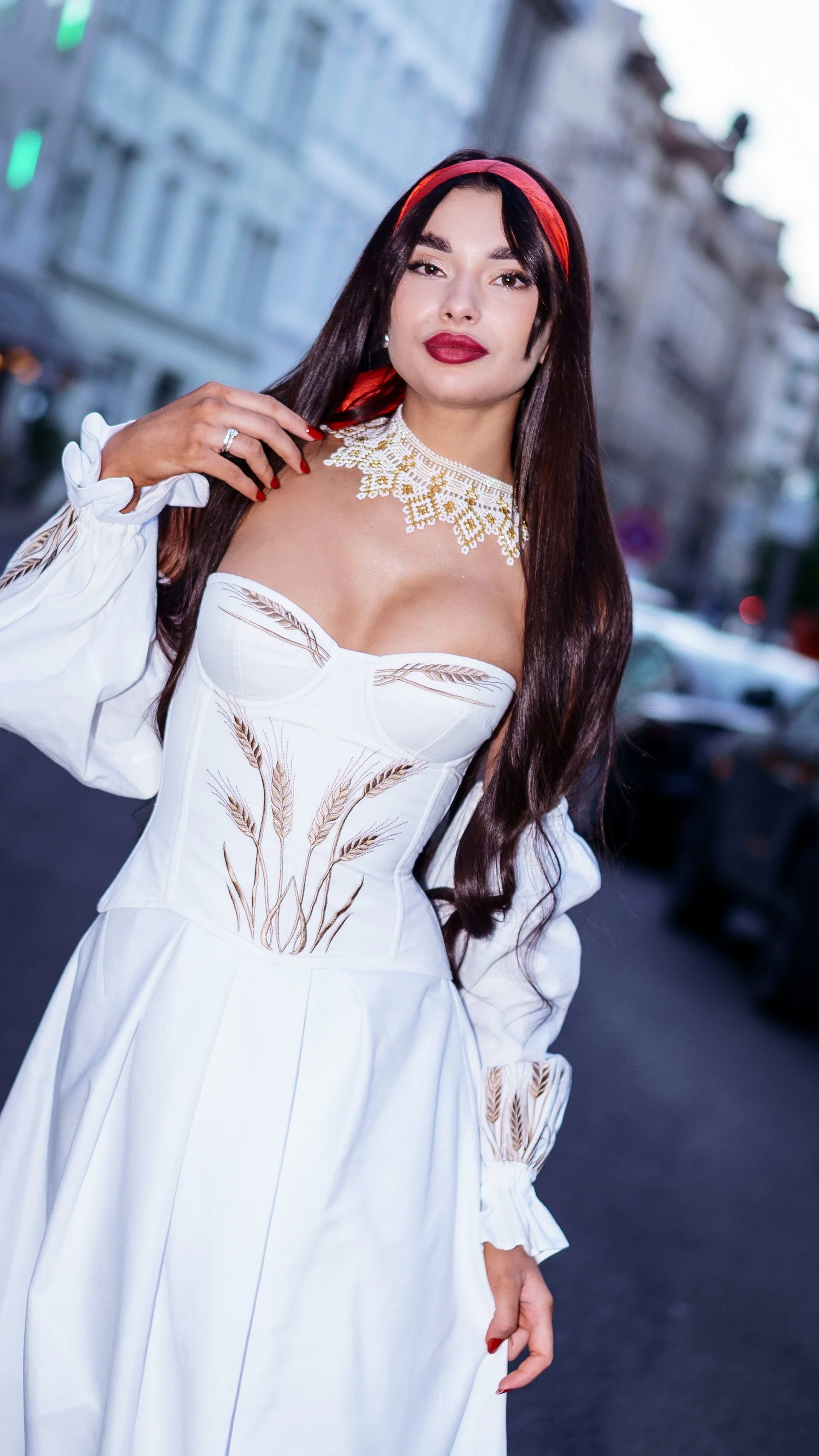 Woman wearing a white dress with embroidered wheat design, a red headband, and a gold and white beaded necklace, standing outdoors on a city street.