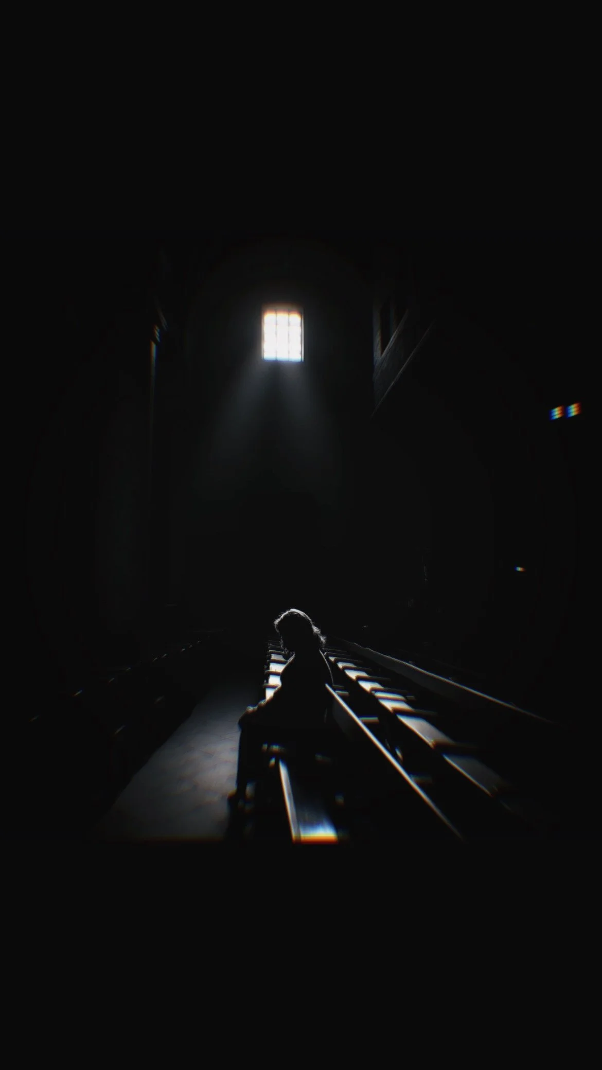Silhouette of a person sitting alone on a church pew inside a dark church with a small window illuminating the space.