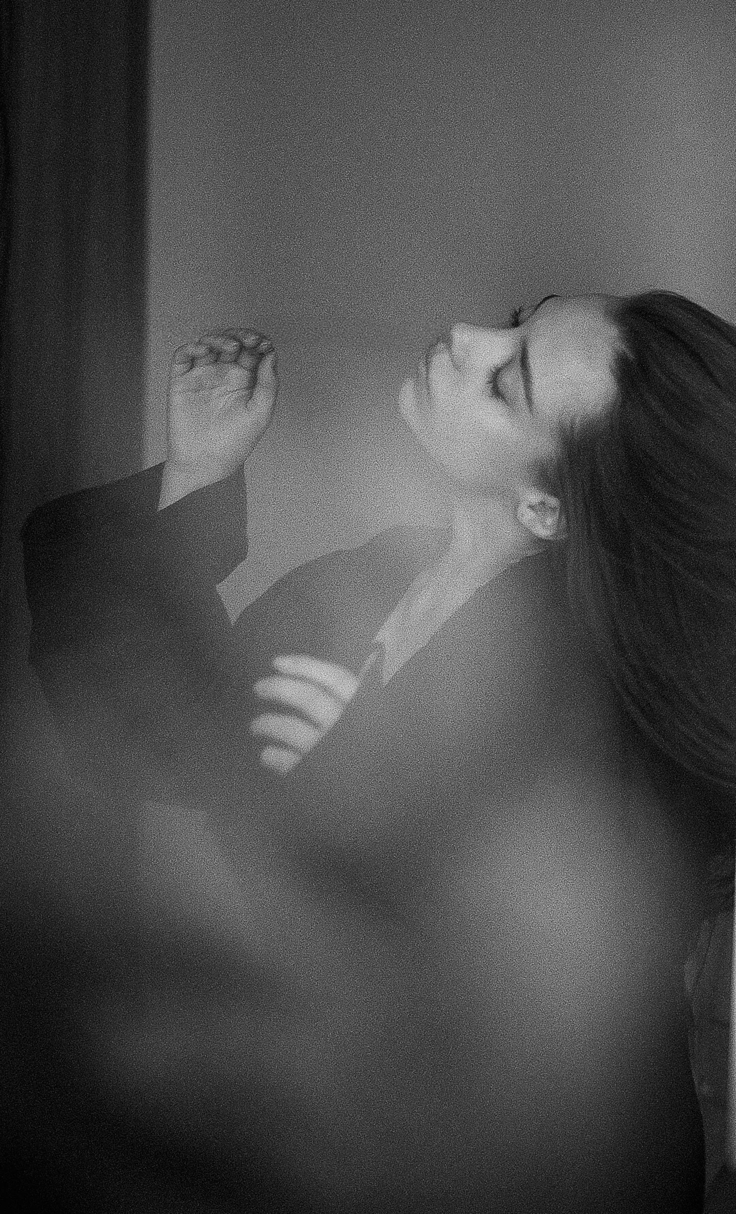 A black and white photo of a woman with long hair, eyes closed, head tilted back, smiling and touching her face with one hand, standing against a plain background.