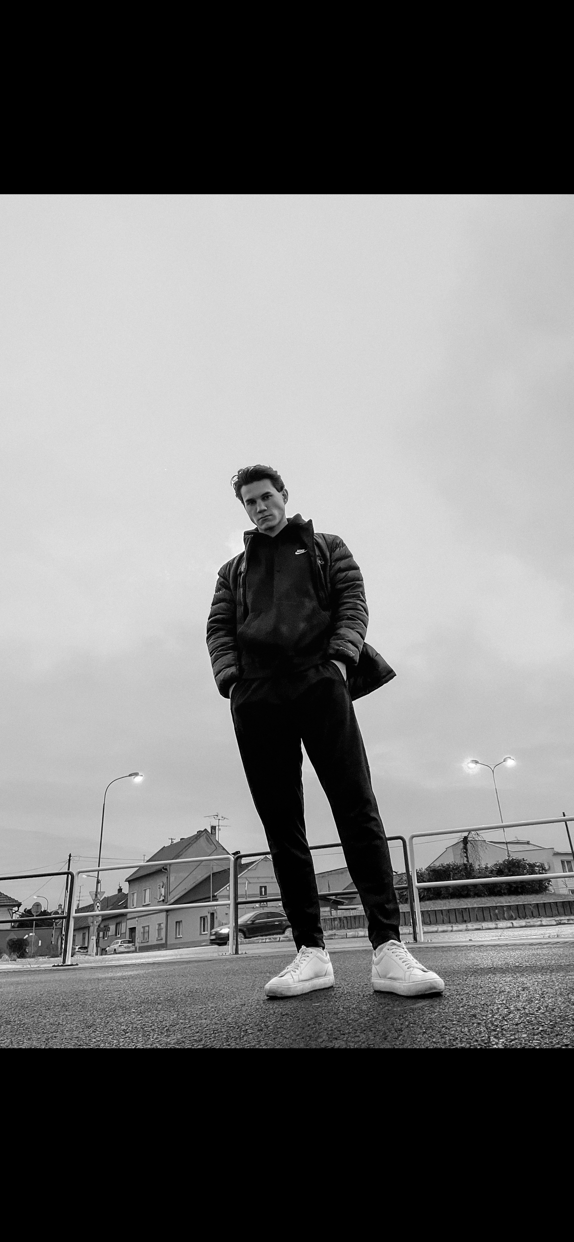 A young man standing outdoors on a cloudy day, wearing a black Nike jacket, black pants, and white sneakers, with houses and streetlights in the background.