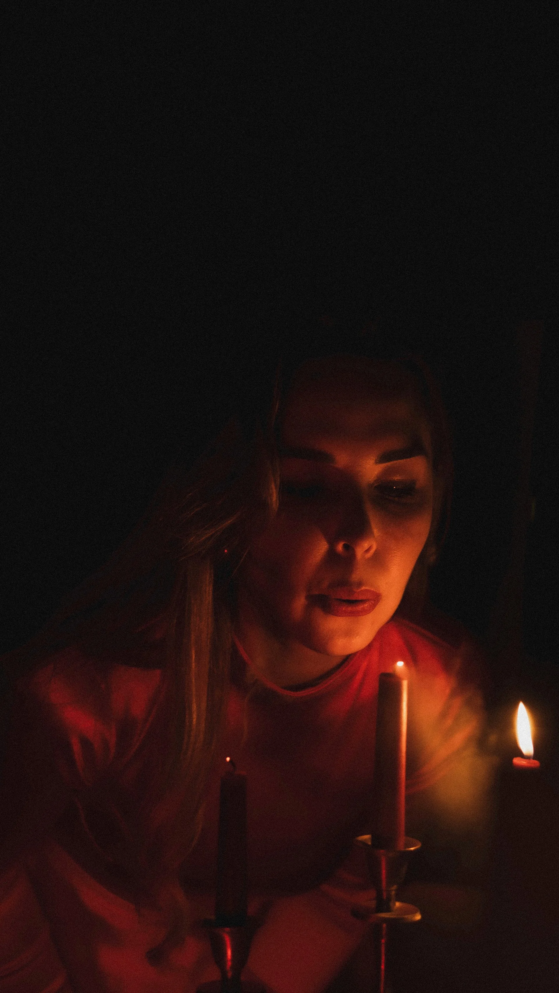 A woman in a red shirt leans in towards candles in a dark room, illuminated by their glow.