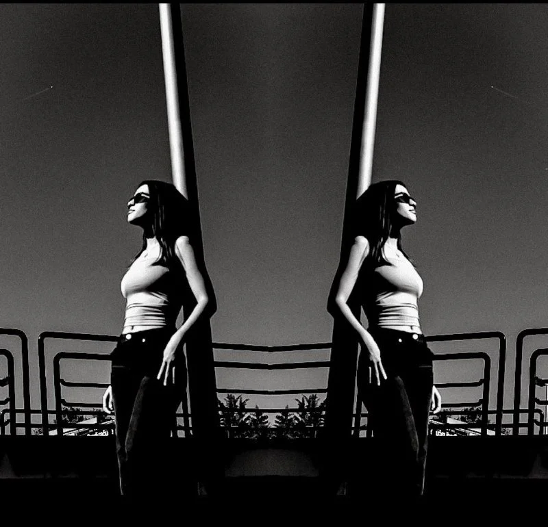 A black-and-white photo of a woman standing on a balcony, looking to the right, with a mirrored reflection creating a symmetrical image.