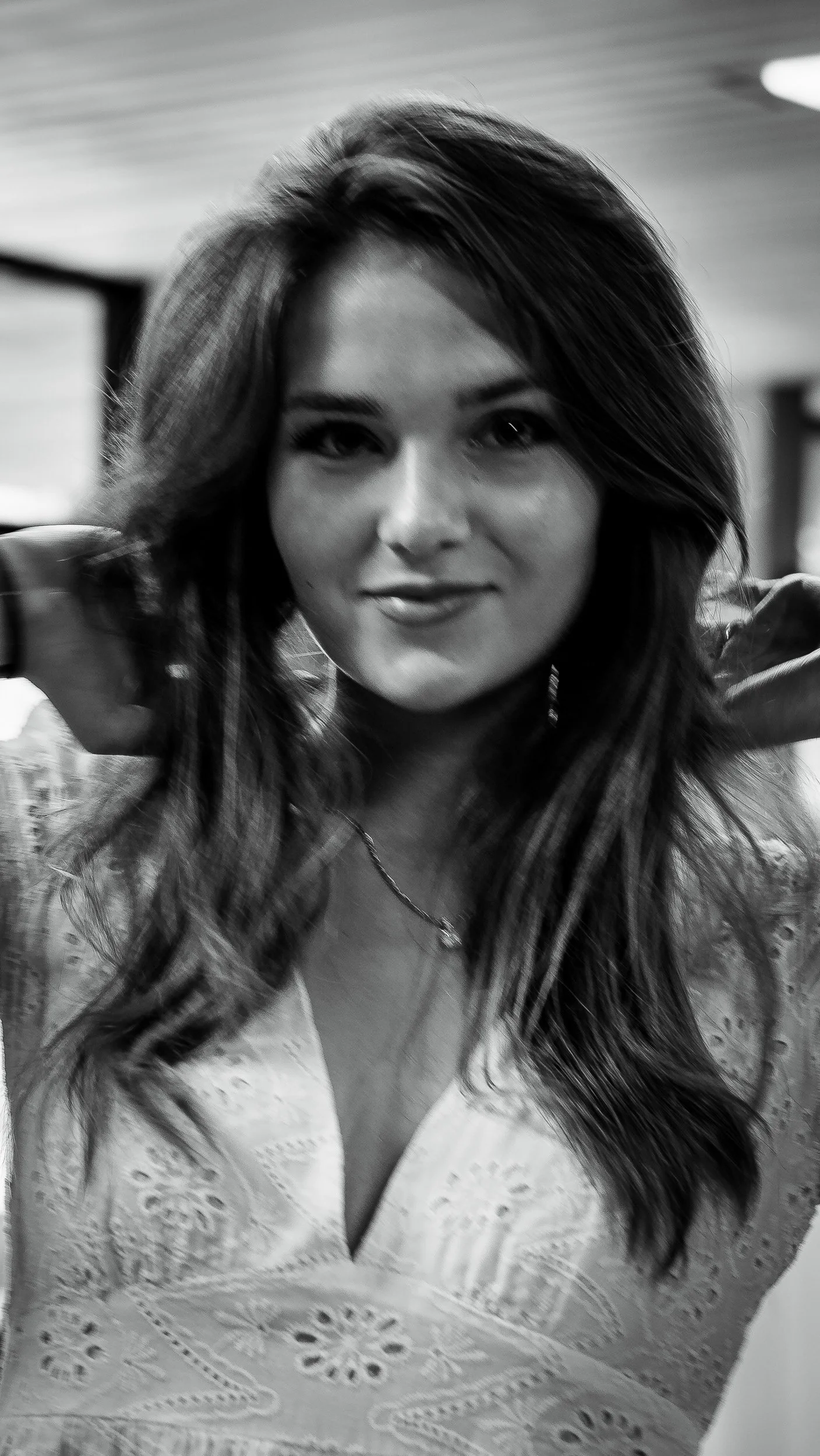 A woman with long wavy hair is adjusting her hair, smiling softly, wearing a light-colored top with intricate lace details and a necklace, in an indoor setting.