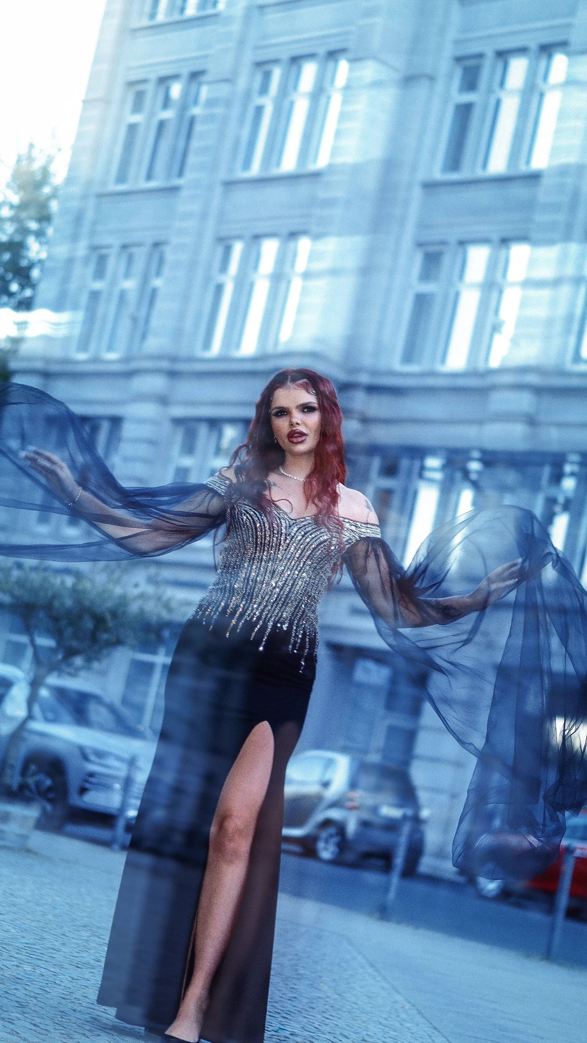 A woman with red hair wearing a glamorous dress with sheer black sleeves and a thigh-high slit, standing outside in an urban setting with buildings and cars reflected in the glass behind her.