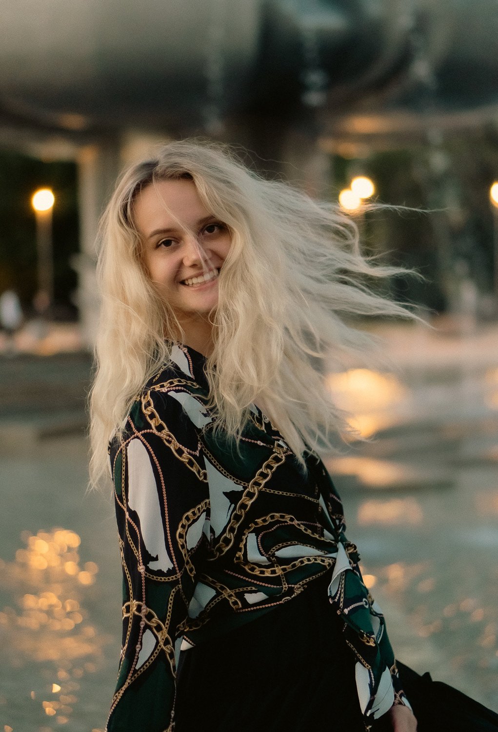 A woman with long, wavy blonde hair smiling outdoors at sunset, wearing a black blouse with a gold chain pattern.