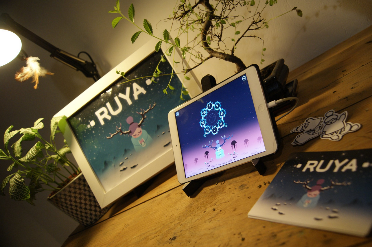 A decorated tabletop features a framed illustrated poster of a reindeer with a red nose, a tablet displaying a similar scene, paper cutouts of reindeers, a potted plant with green leaves, and a brochure or booklet with the word 'RUYA' on the cover.