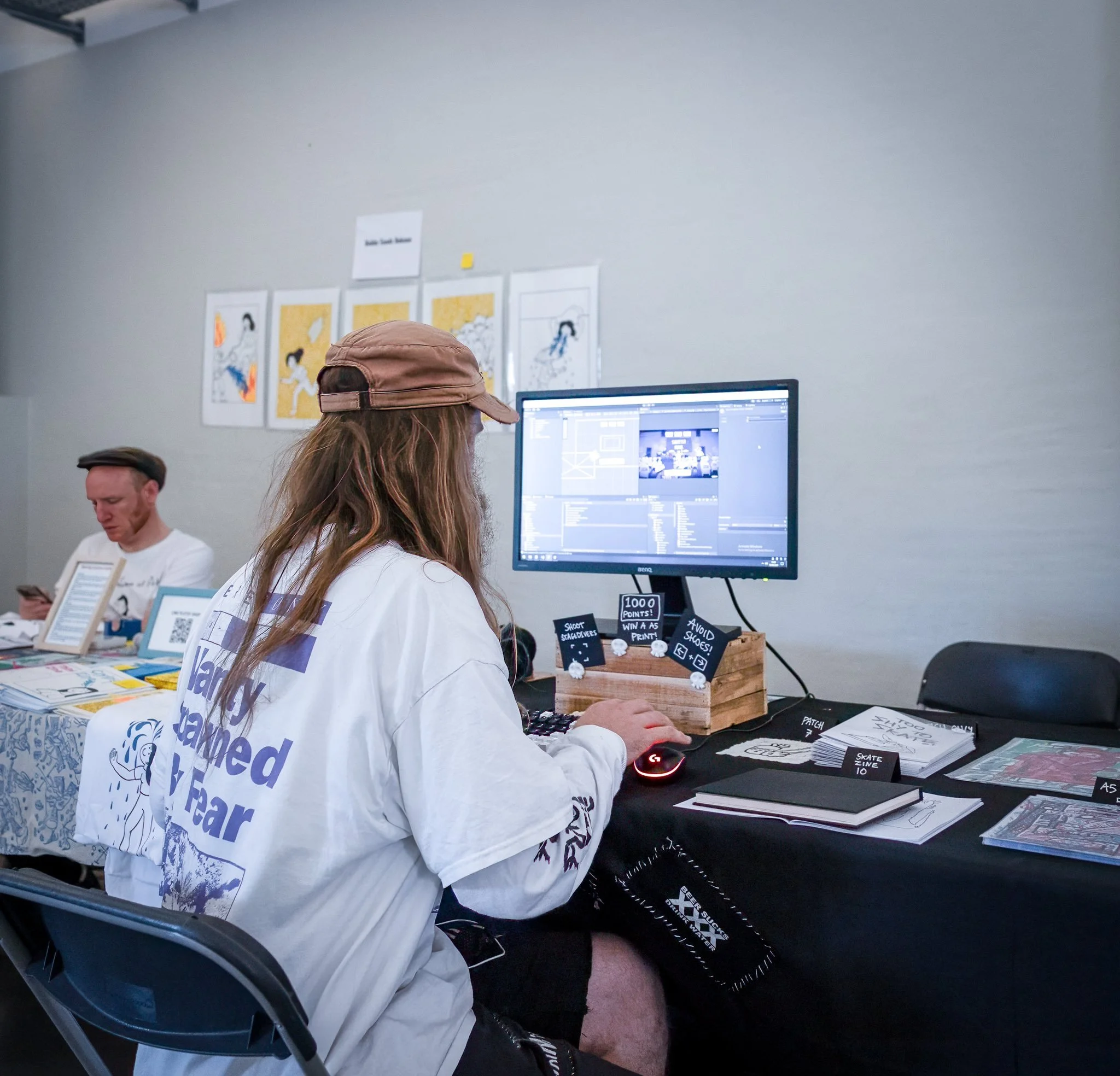 A person with long hair, wearing a beige cap and a white shirt, sitting at a table working on a computer with a large monitor in a room. There is another person in the background, wearing a hat and looking at a phone. The table has various papers, books, and small signs, and the wall behind has framed art.