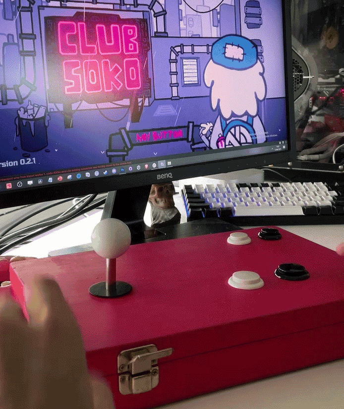 A gaming setup with a red arcade stick featuring an eight-directional joystick and four buttons, positioned in front of a computer monitor displaying an animated scene of a character with blue hair and a pink neon sign that reads 'CLUB SOKO'.