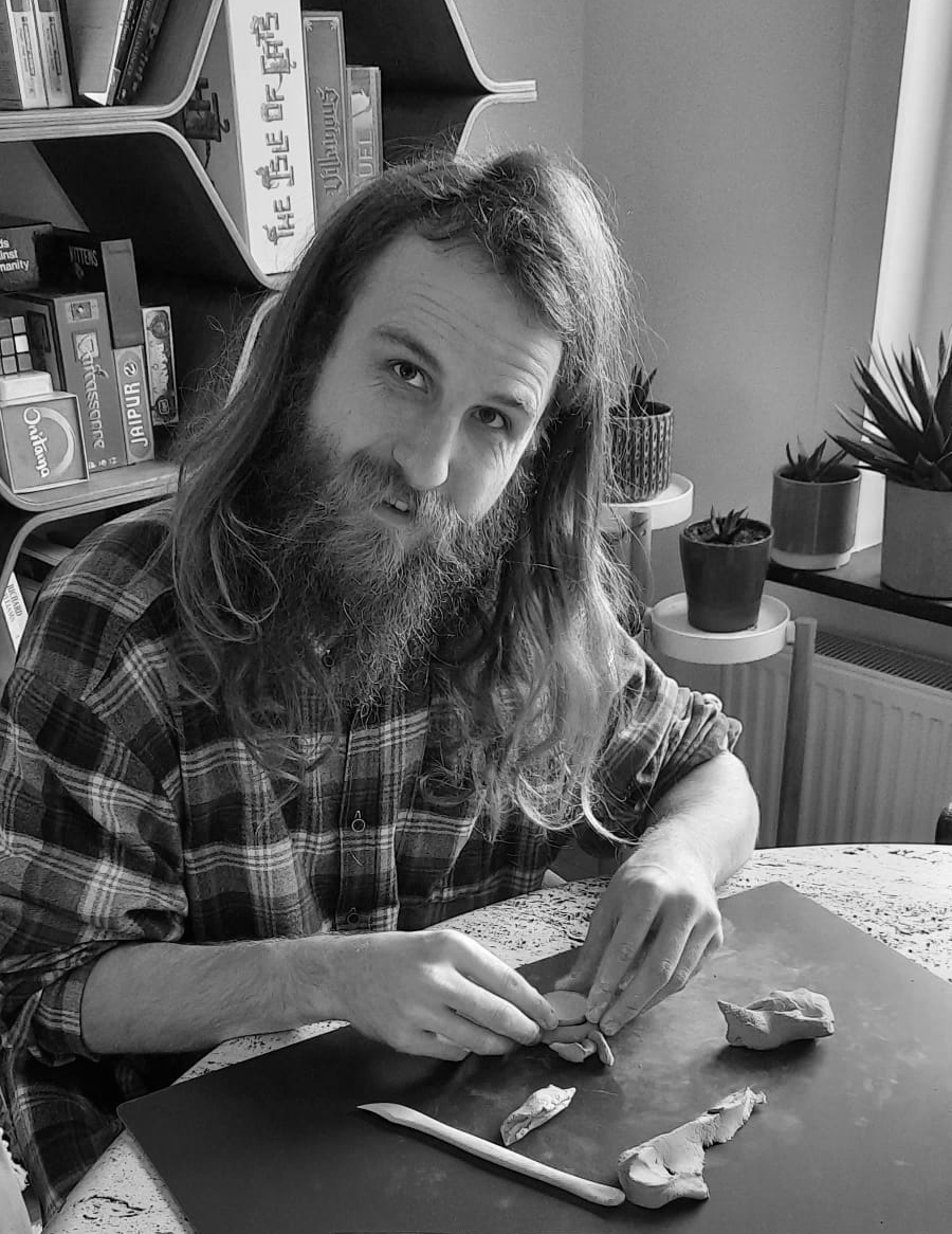 A man with long hair and a beard, wearing a plaid shirt, sitting at a table and shaping clay. Books and potted plants are on shelves and windowsill behind him.