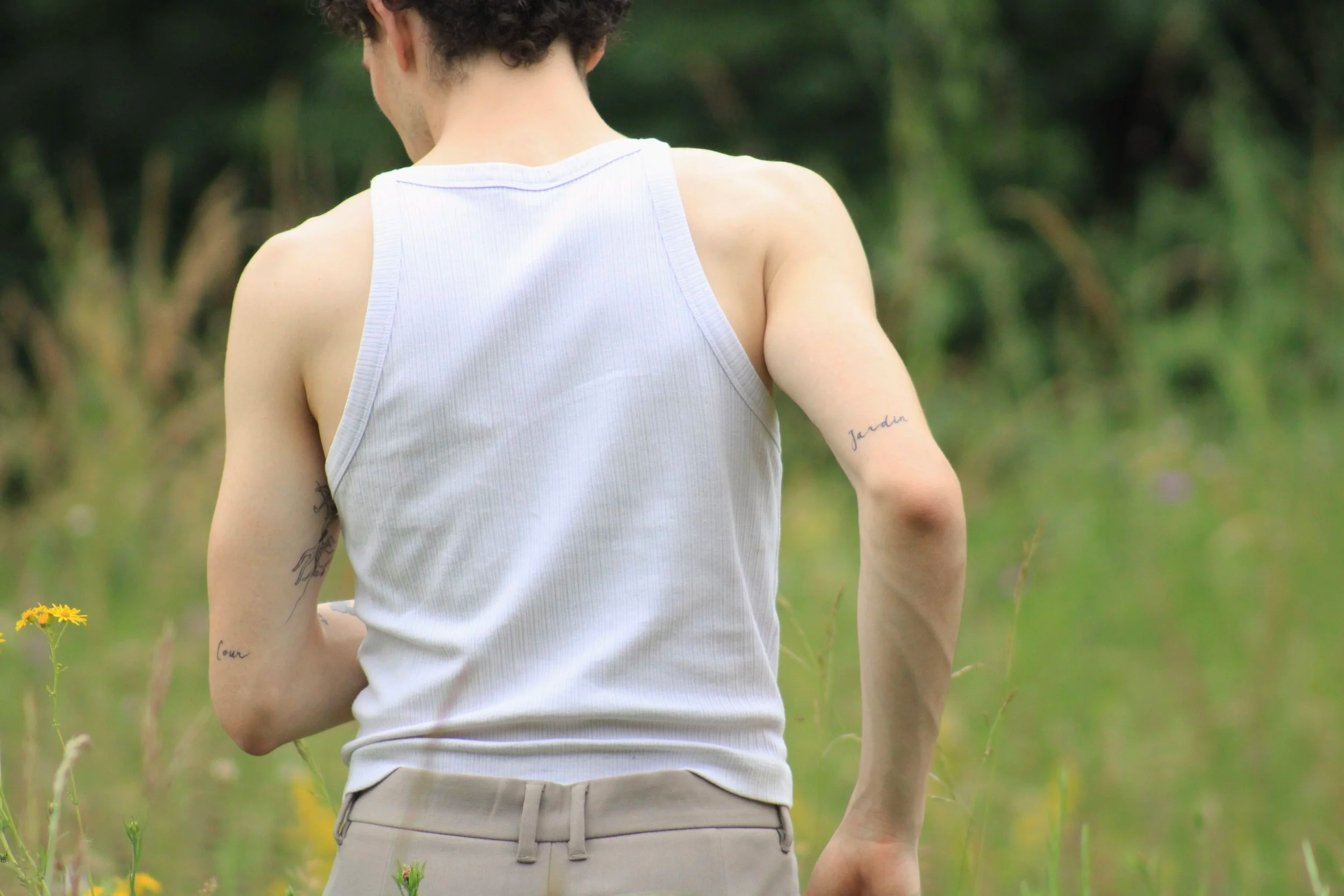 A person wearing a white tank top and beige pants standing in a lush green outdoor area, seen from behind, with visible tattoos on the arms.