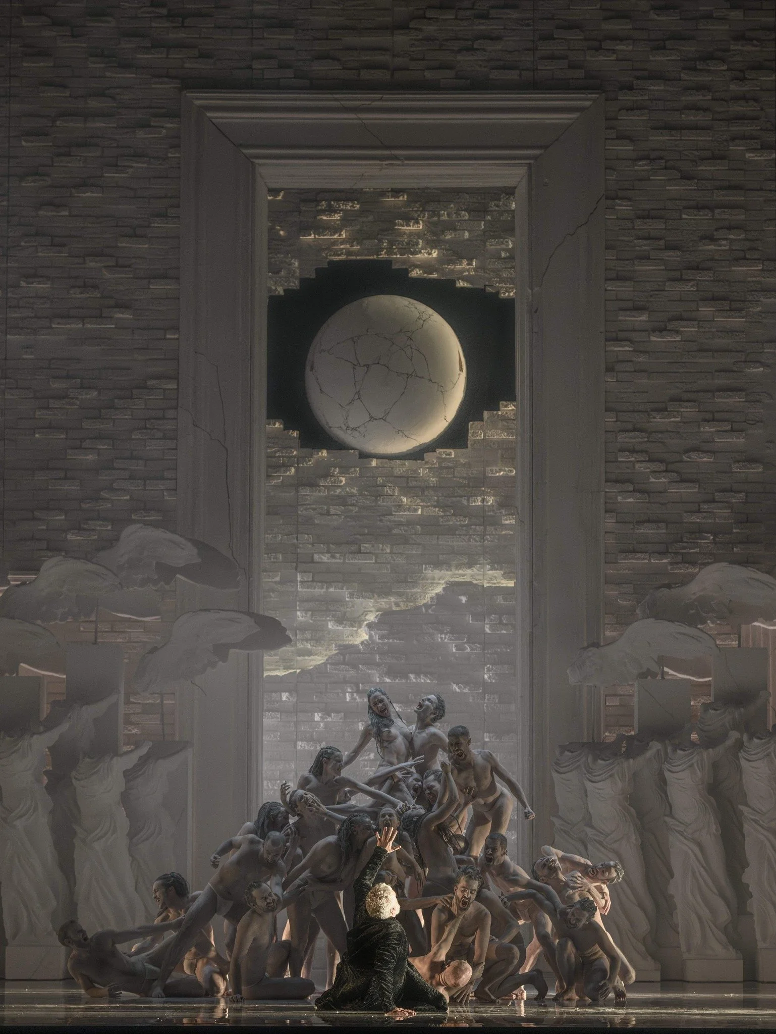 A stage scene with a woman in black and white dress surrounded by multiple nude dancers in various dramatic poses. The background features a tall brick wall with a broken frame revealing a moonlit sky and clouds.