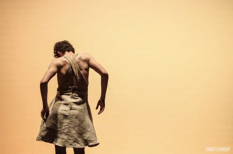 A shirtless person with short curly hair, wearing a beige skirt, standing in front of a plain light yellow background.