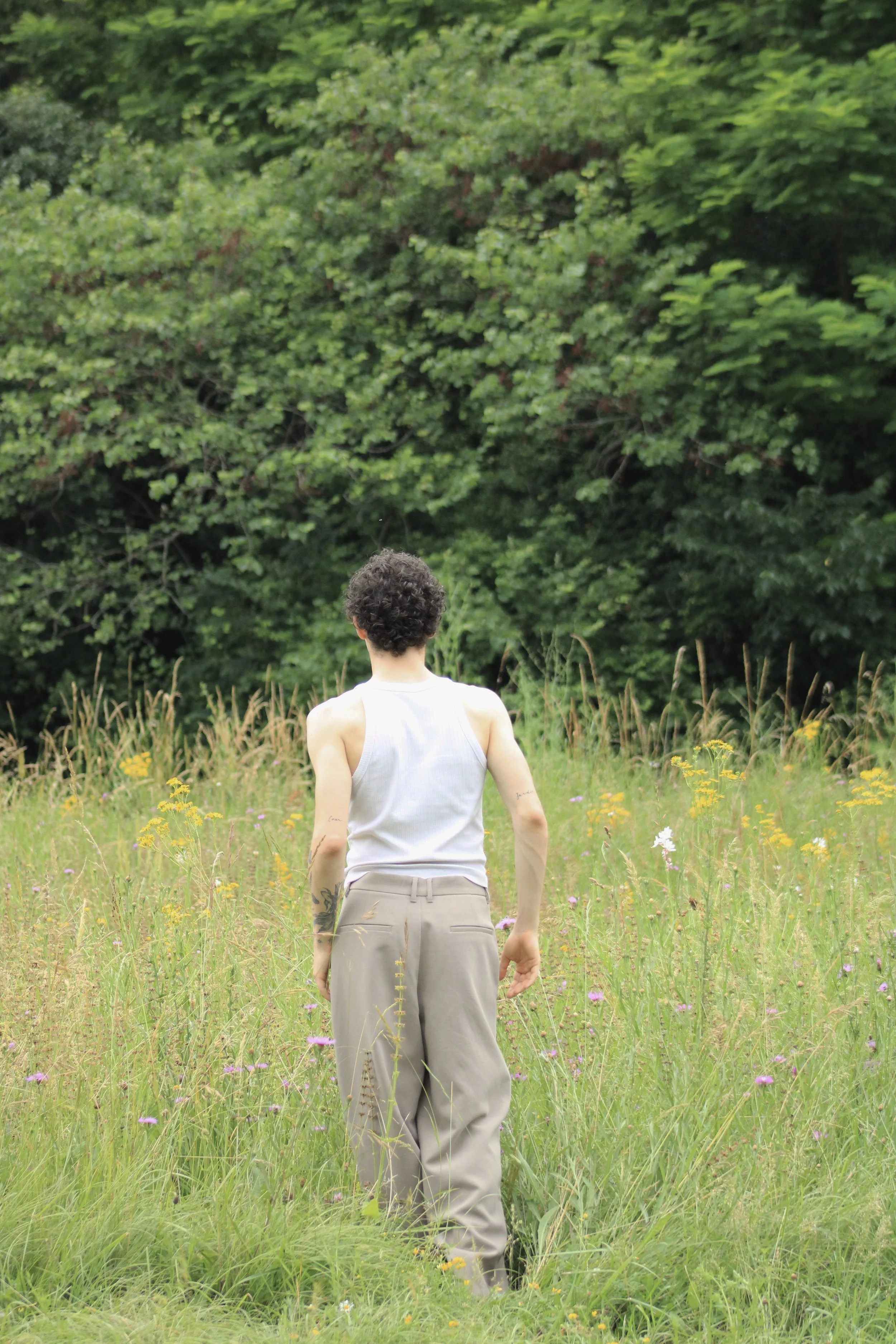 Person with short curly hair and tattoos on arms walking through a grassy meadow with wildflowers, facing away from the camera, with a dense green tree line in the background.