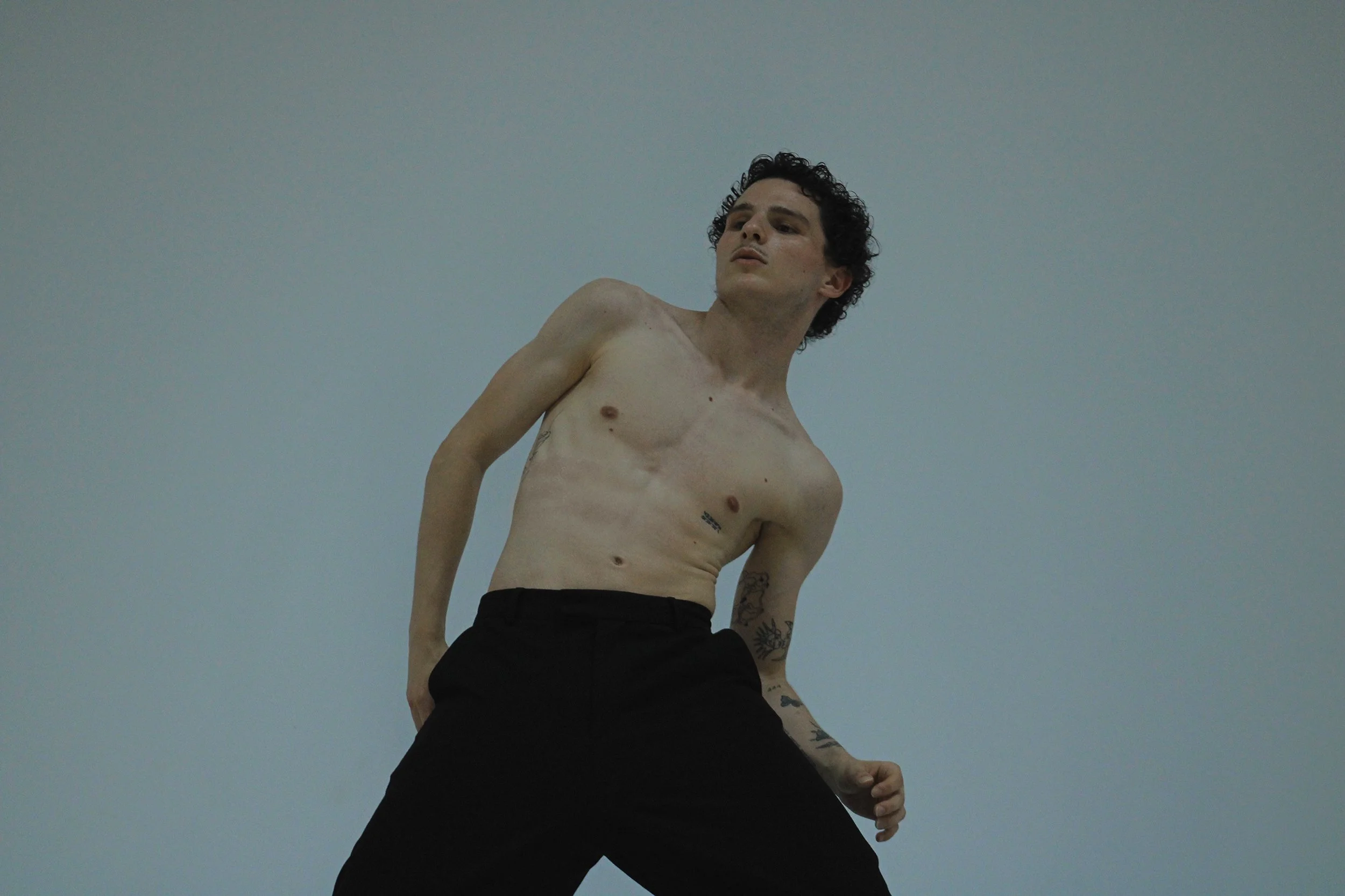 A shirtless man with curly dark hair and tattoos on his left arm and chest, wearing black pants, standing against a plain blue background, looking to the side.