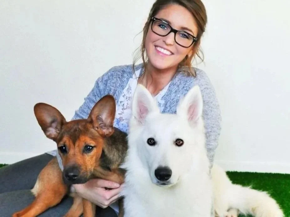 Une femme souriante portant des lunettes est assise sur un tapis vert, accompagnée de deux chiens, un chien blanc et un chien brun, tous deux regardant vers la caméra.