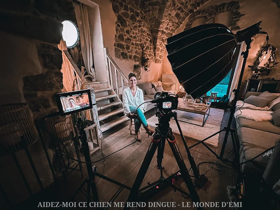 Une femme assise avec un sourire, interviewée ou filmée dans un intérieur avec murs en pierre, fauteuils, et éclairage professionnel, en train de parler au caméra. Il y a un éclairage, un fond avec canapé et éléments décoratifs.