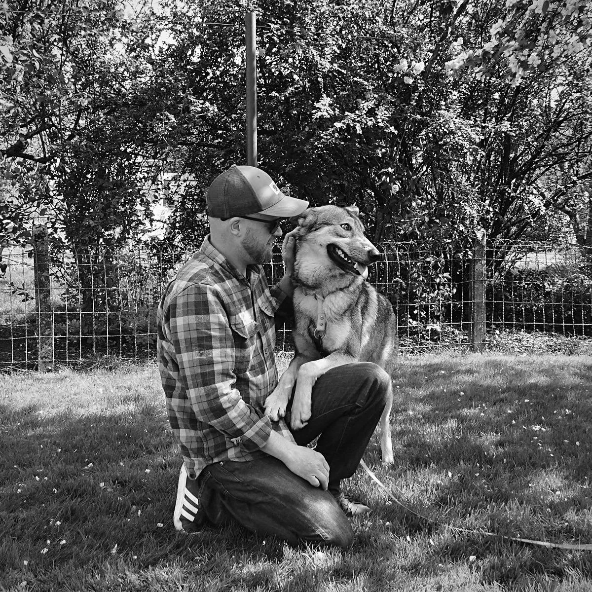 Un homme portant une casquette, lunettes et chemise à carreaux, assis dans l'herbe, caresse un chien de race husky, devant un mur en grillage et des arbres feuillus.
